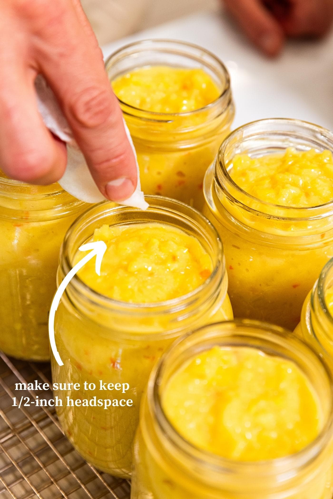 Hand wiping off rim of glass canning jar filled with yellow colored hot pepper mustard showing the headspace at the top.