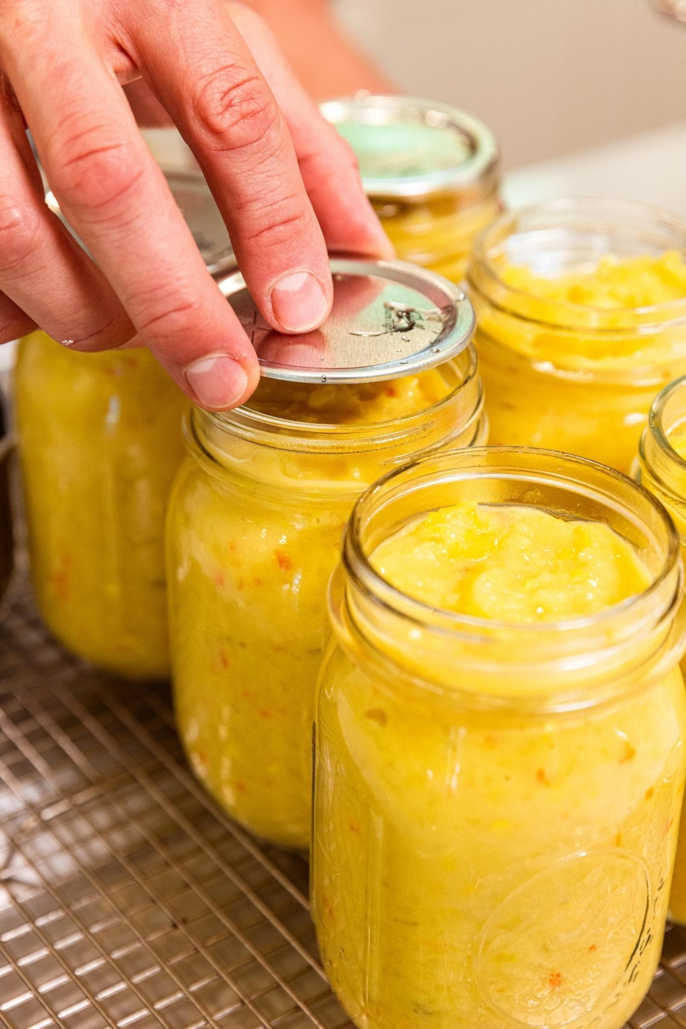 Hand placing lid on glass canning jar filled with yellow colored hot pepper mustard with other jars around.