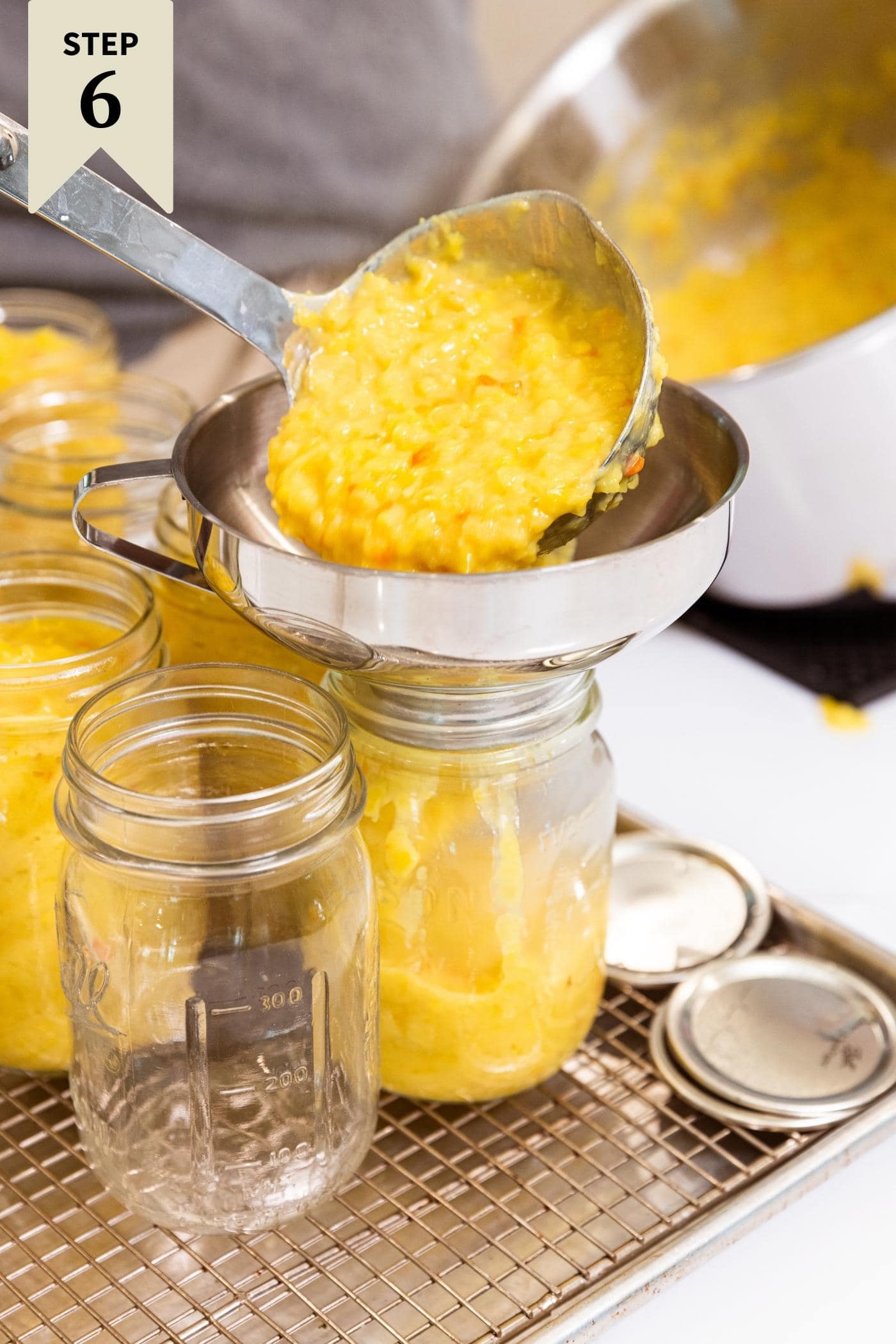 Hand holding ladle putting hot pepper mustard into glass jars sitting on wire rack with large metal stockpot in background.