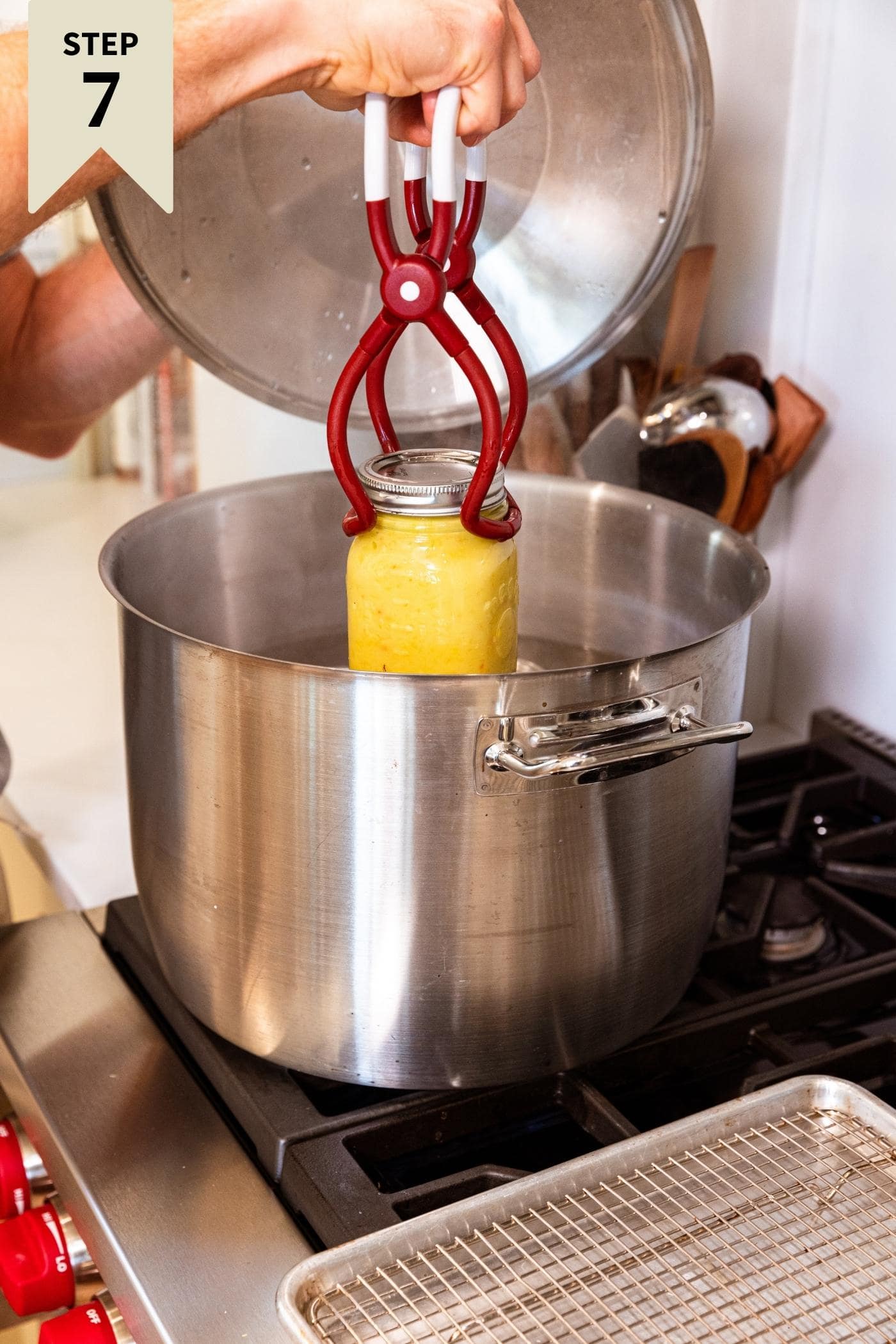 Hand putting glass jar filled with hot pepper mustard into water bath canner using red plastic clamps.