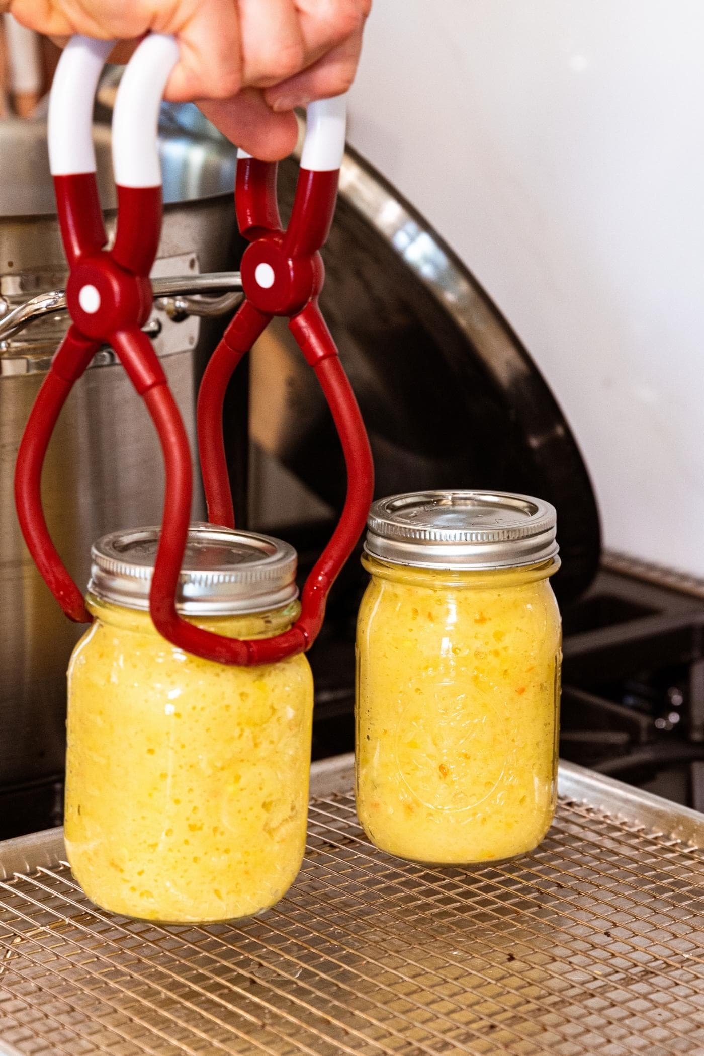 Red plastic tongs placing glass jar filled with yellow-colored hot pepper mustard onto metal cooling rack after being removed from water bath canner on stove.