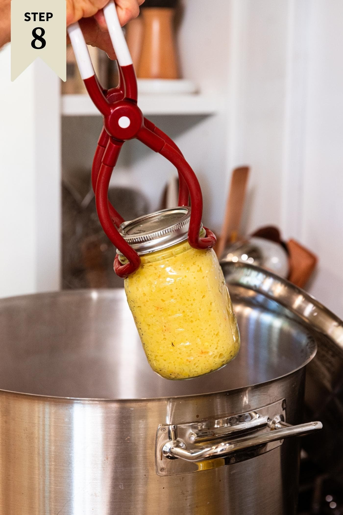 Hand removing glass canning jar filled with yellow hot pepper mustard from water bath canner on stovetop.