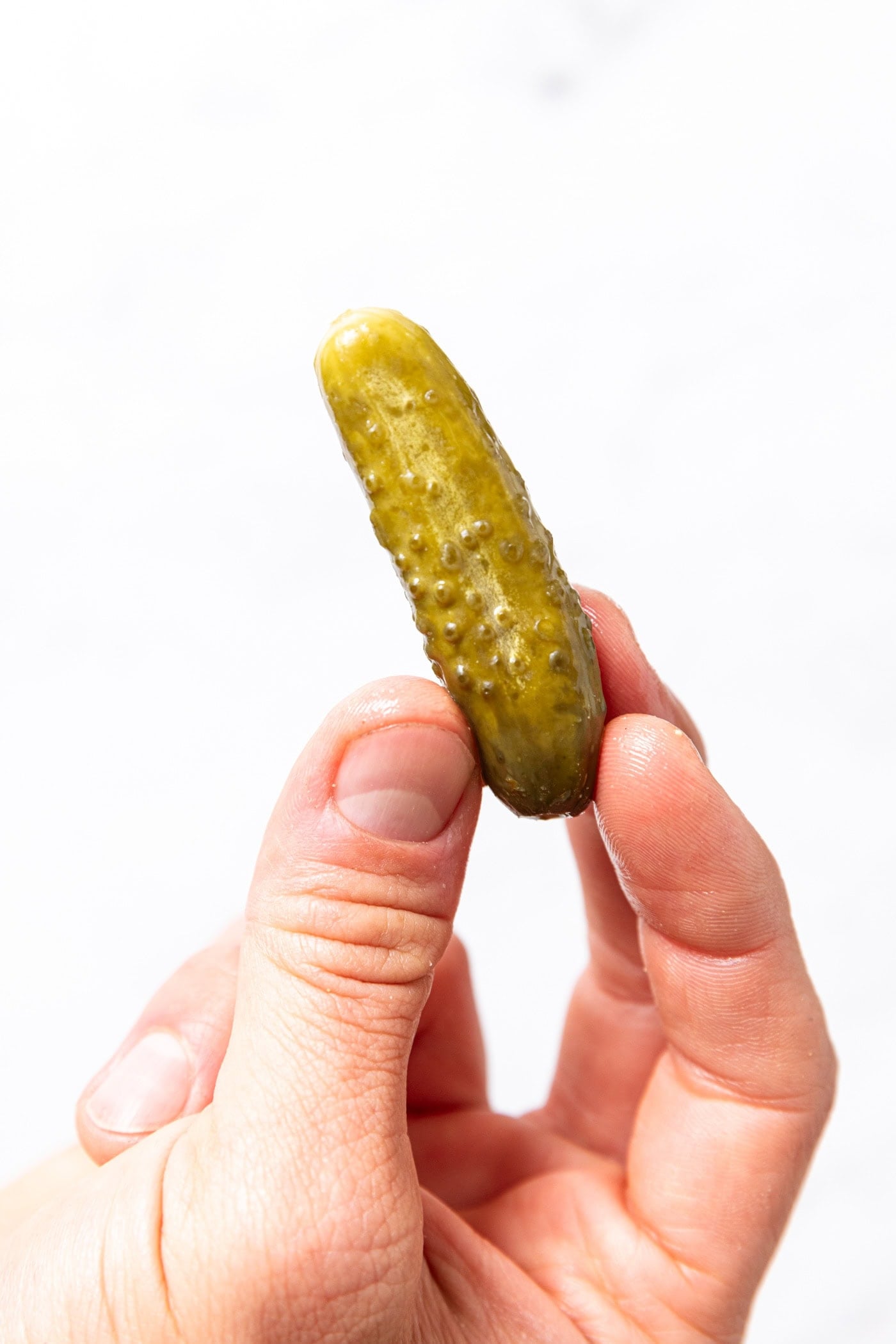 Hand holding mustard pickle with yellowish exterior above white surface.
