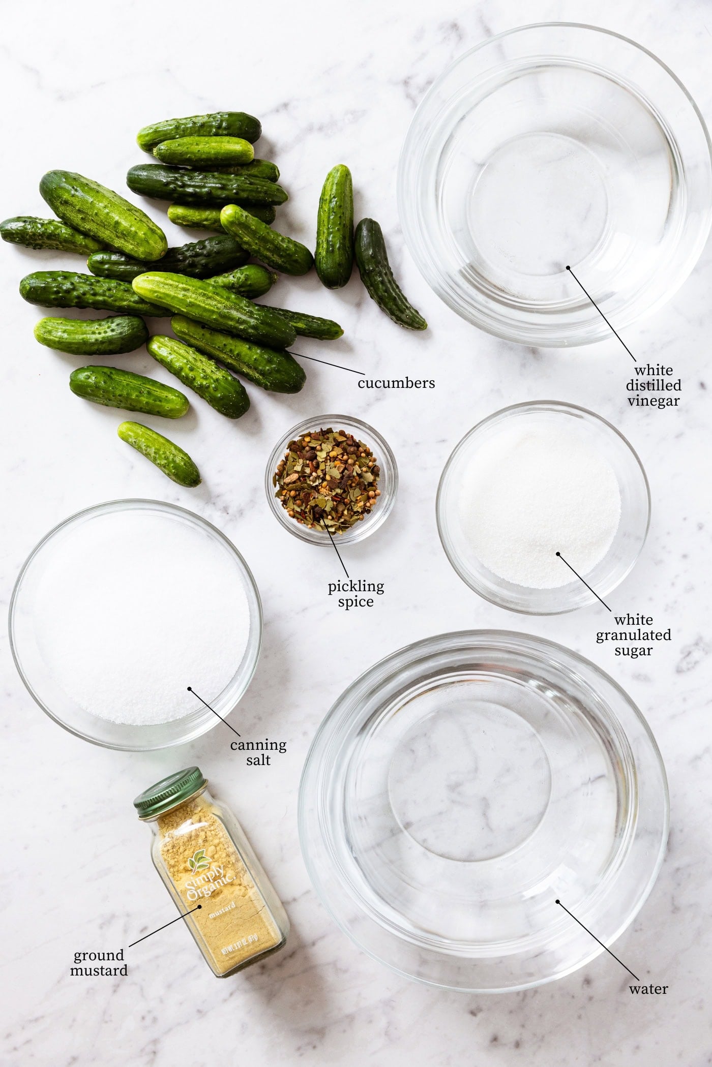 White marble surface filled with all ingredients needed to make mustard pickles including raw cucumbers, vinegar, water, sugar, ground mustard, and more.