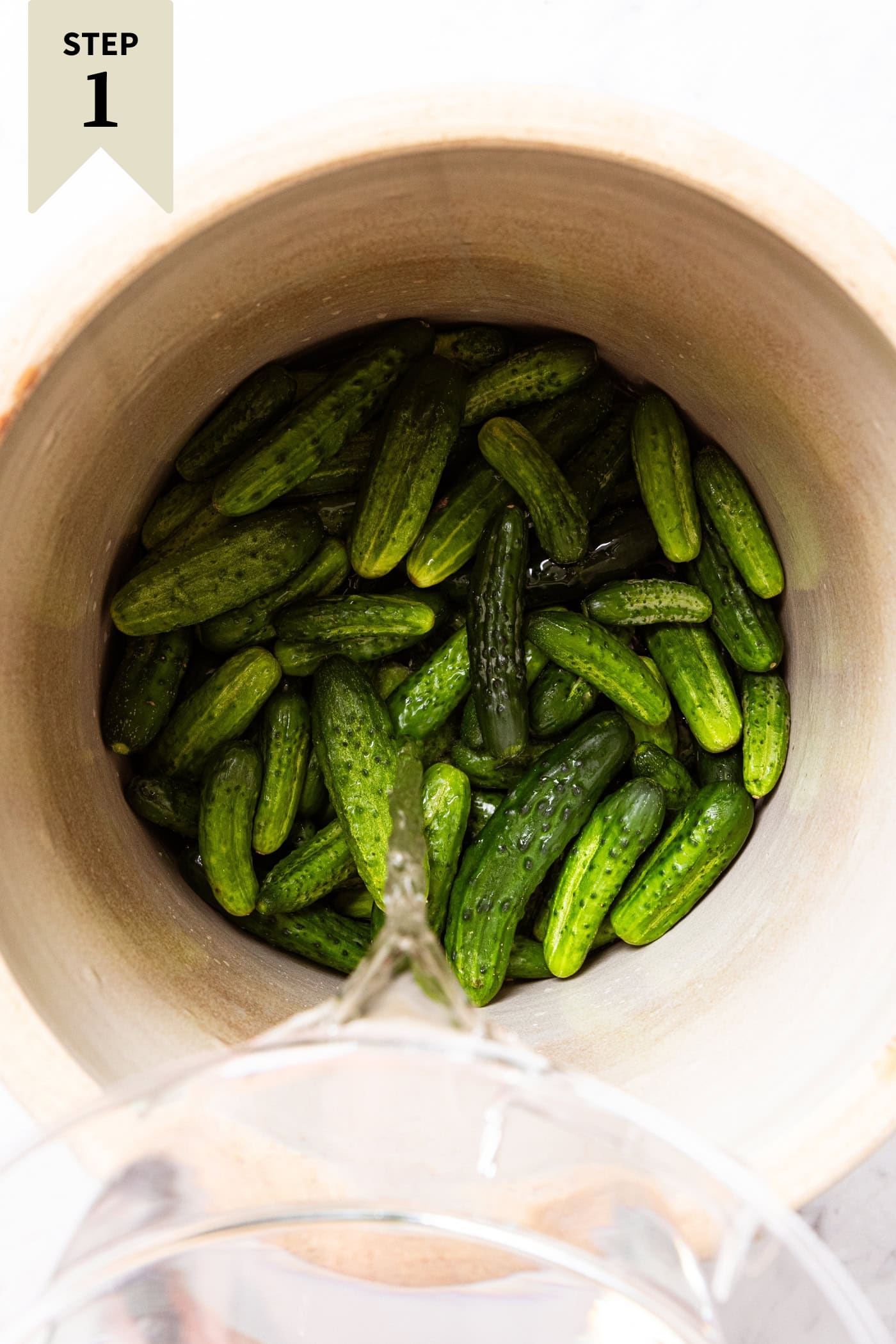 Cucumbers in a large crock with water being poured on top to soak.