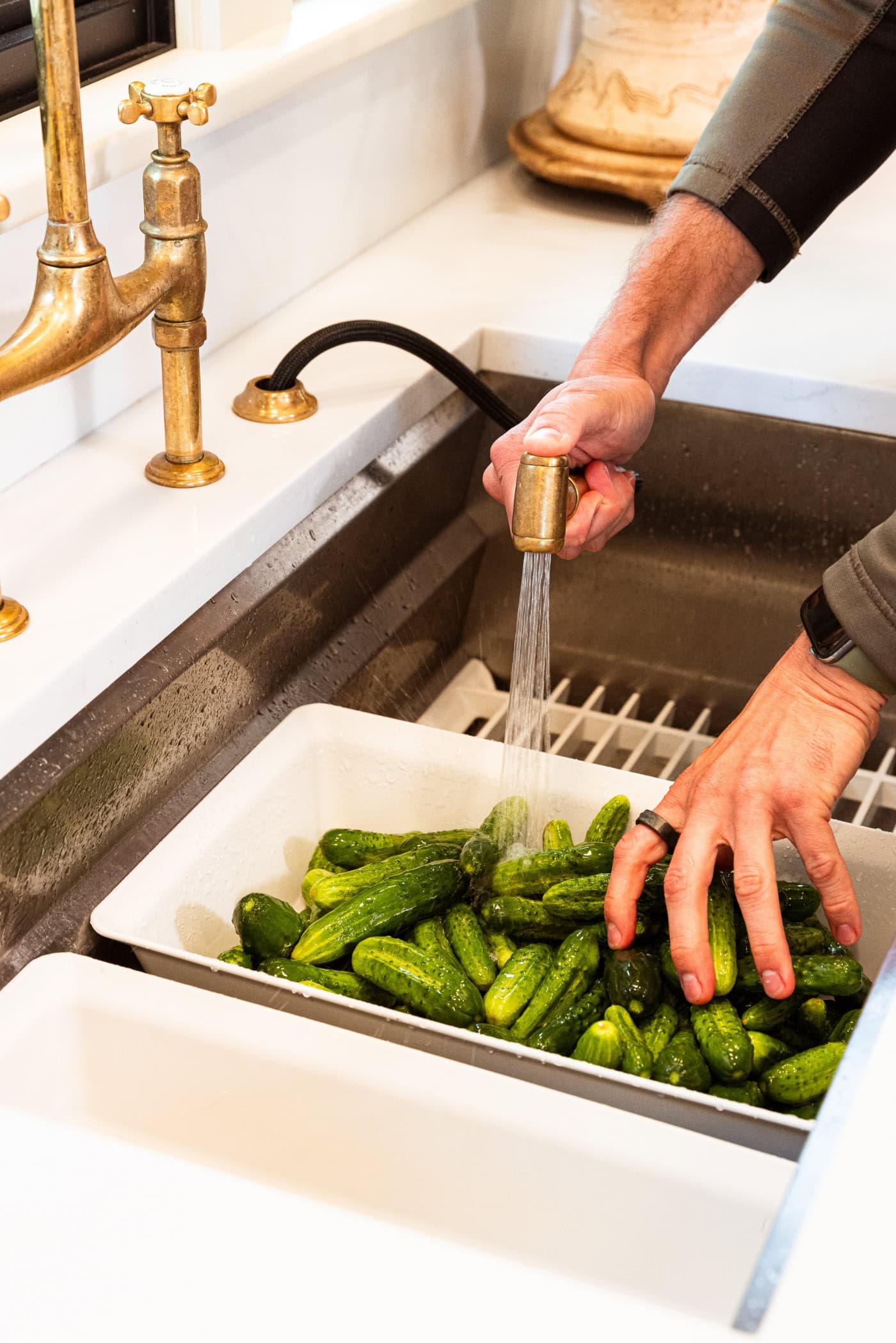 Hand using faucet sprayer to spray off cucumbers in a white colander.