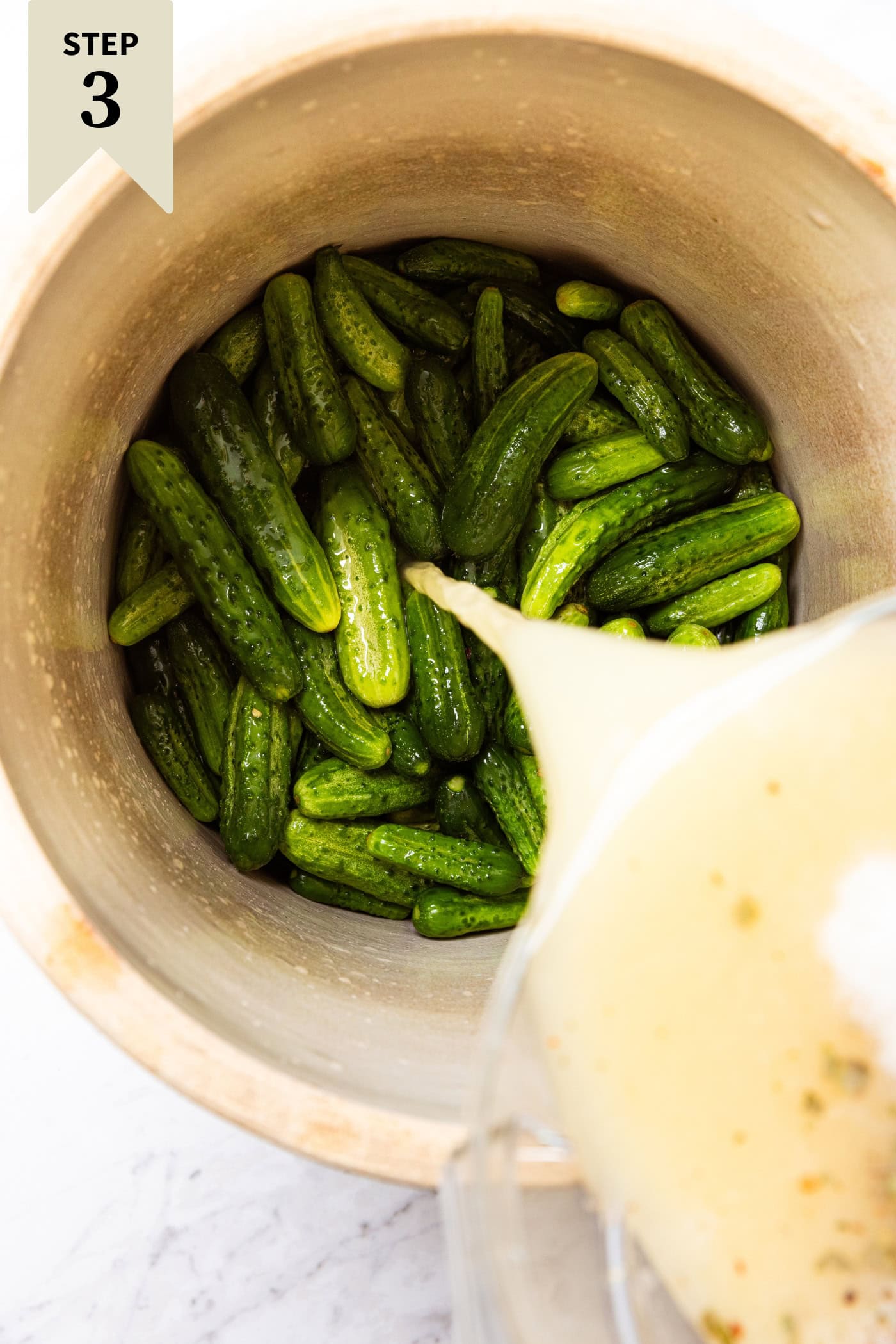 Brine being poured on top of soaked cucumbers in a crock to make mustard pickles.