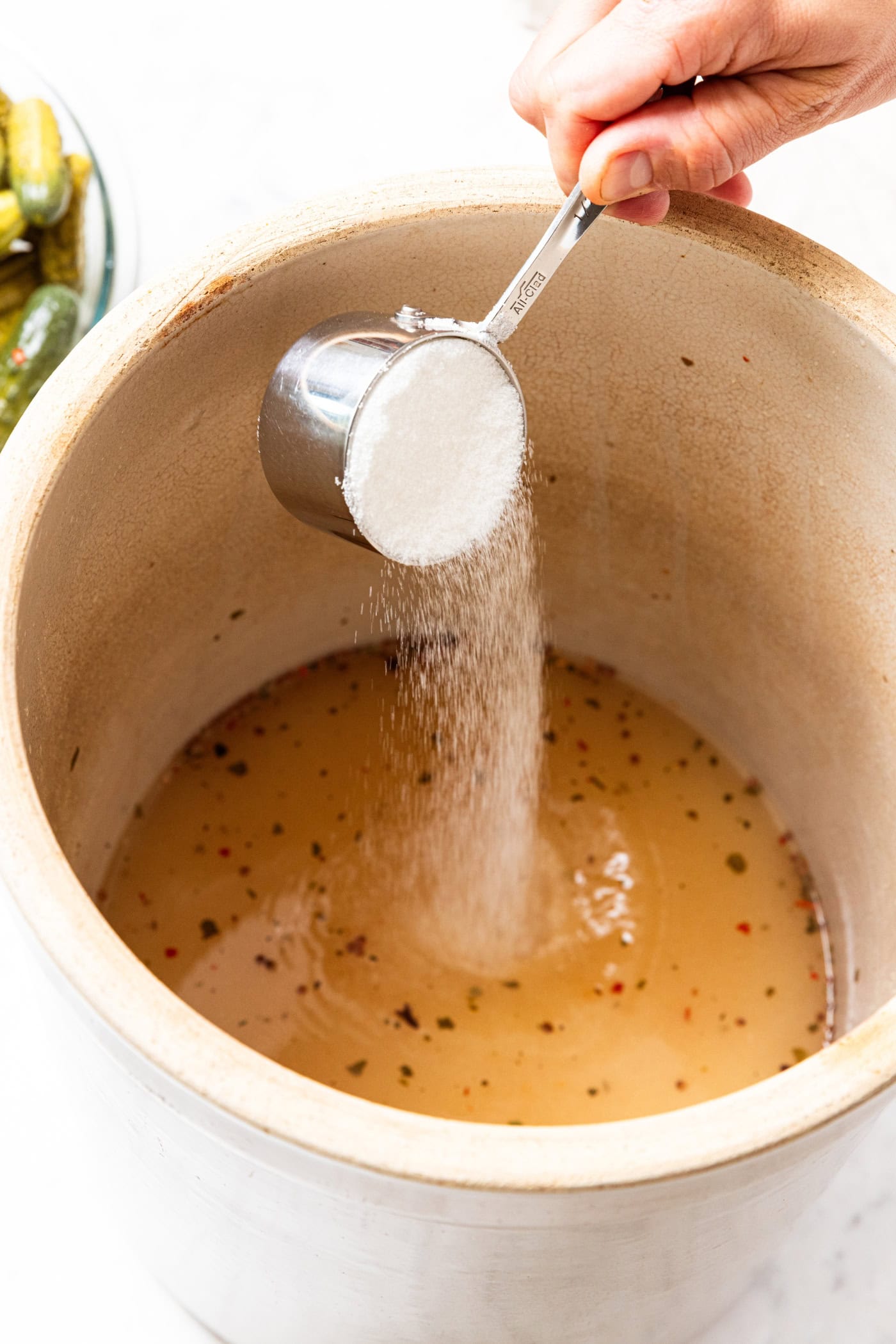 Measuring cup sprinkling sugar into a large crock with pickling brine.