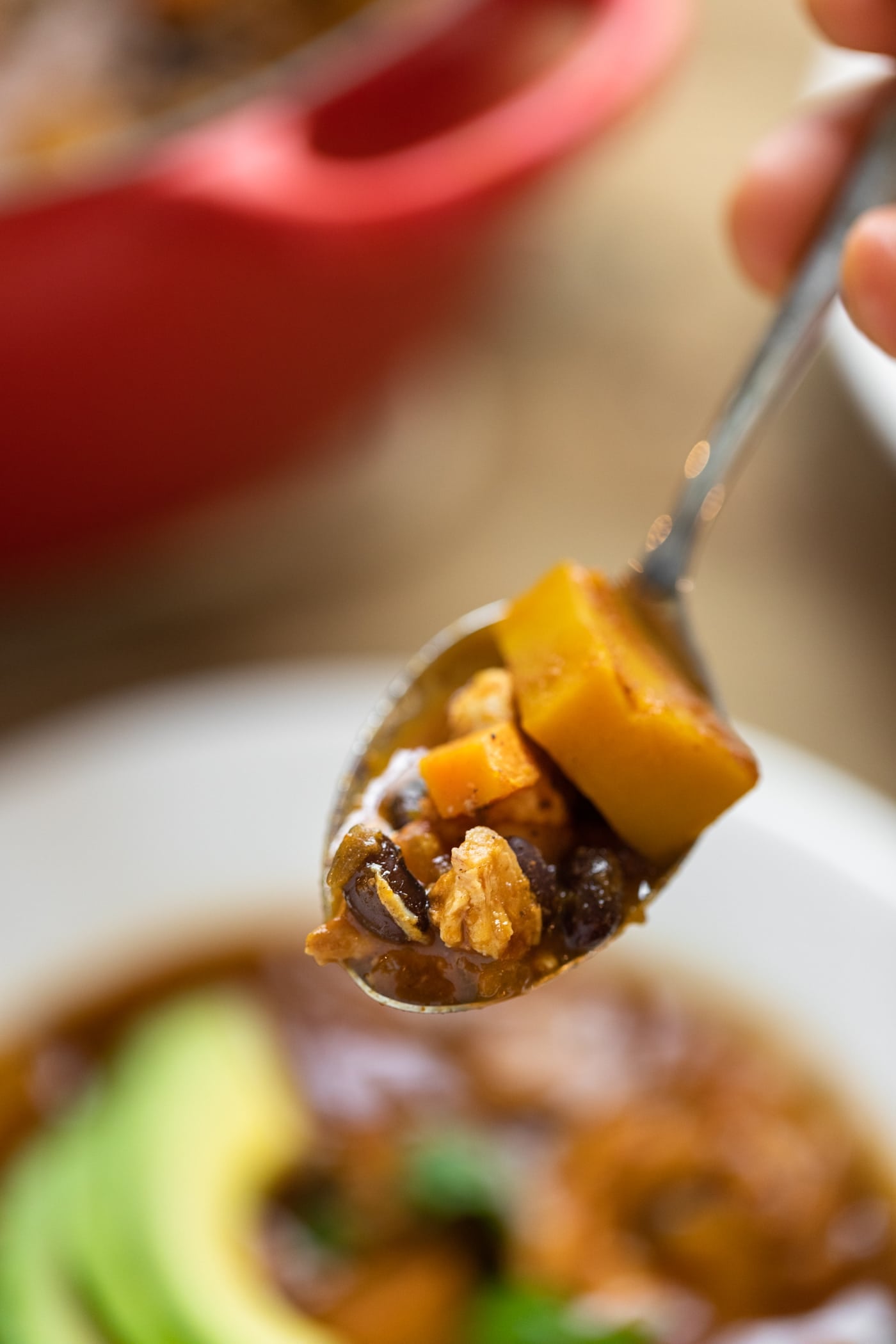Spoon holding bite full of chili containing black beans and squash with bowl in background