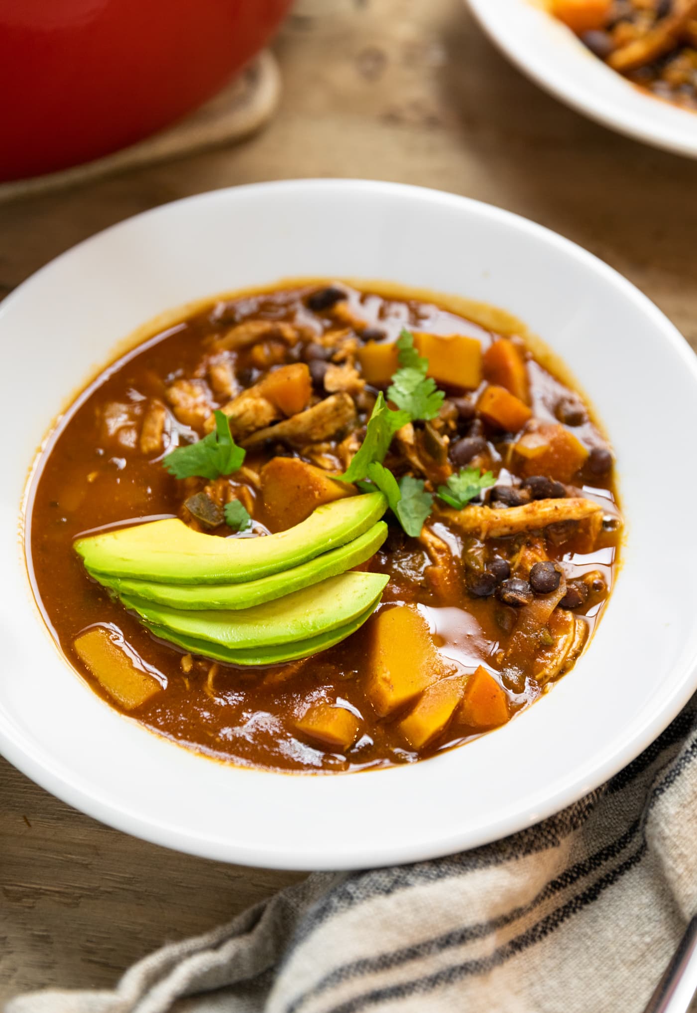 White ceramic bowl filled with red color chili with pieces of butternut squash and chicken with slices of avocado on top with more soup in background