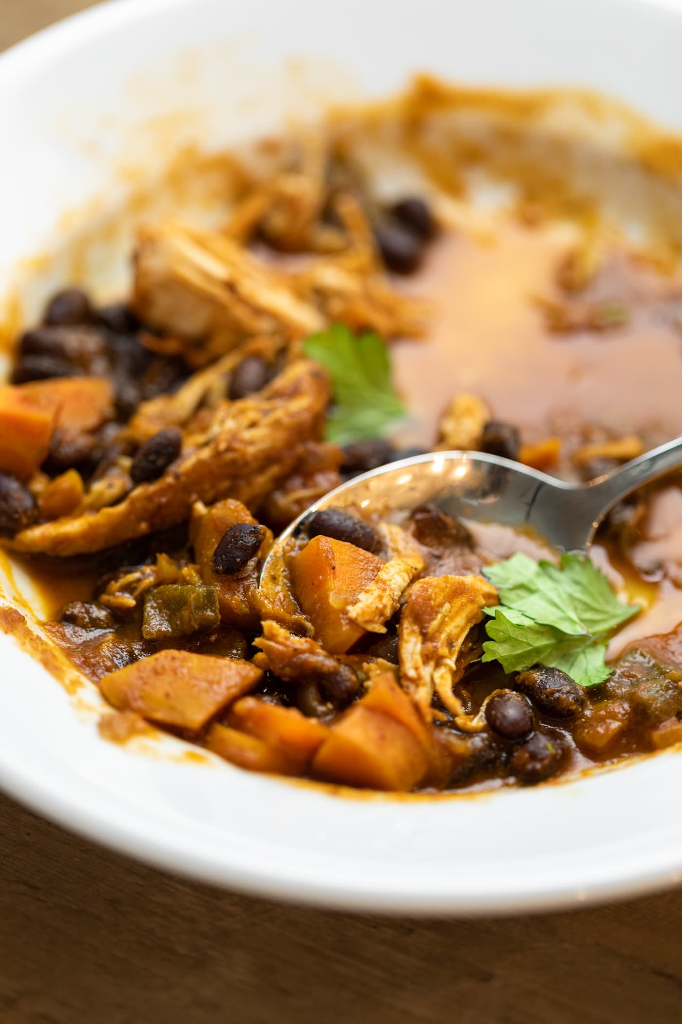 Spoon scooping bite full of chili with squash and chicken sitting in a half-eaten white bowl