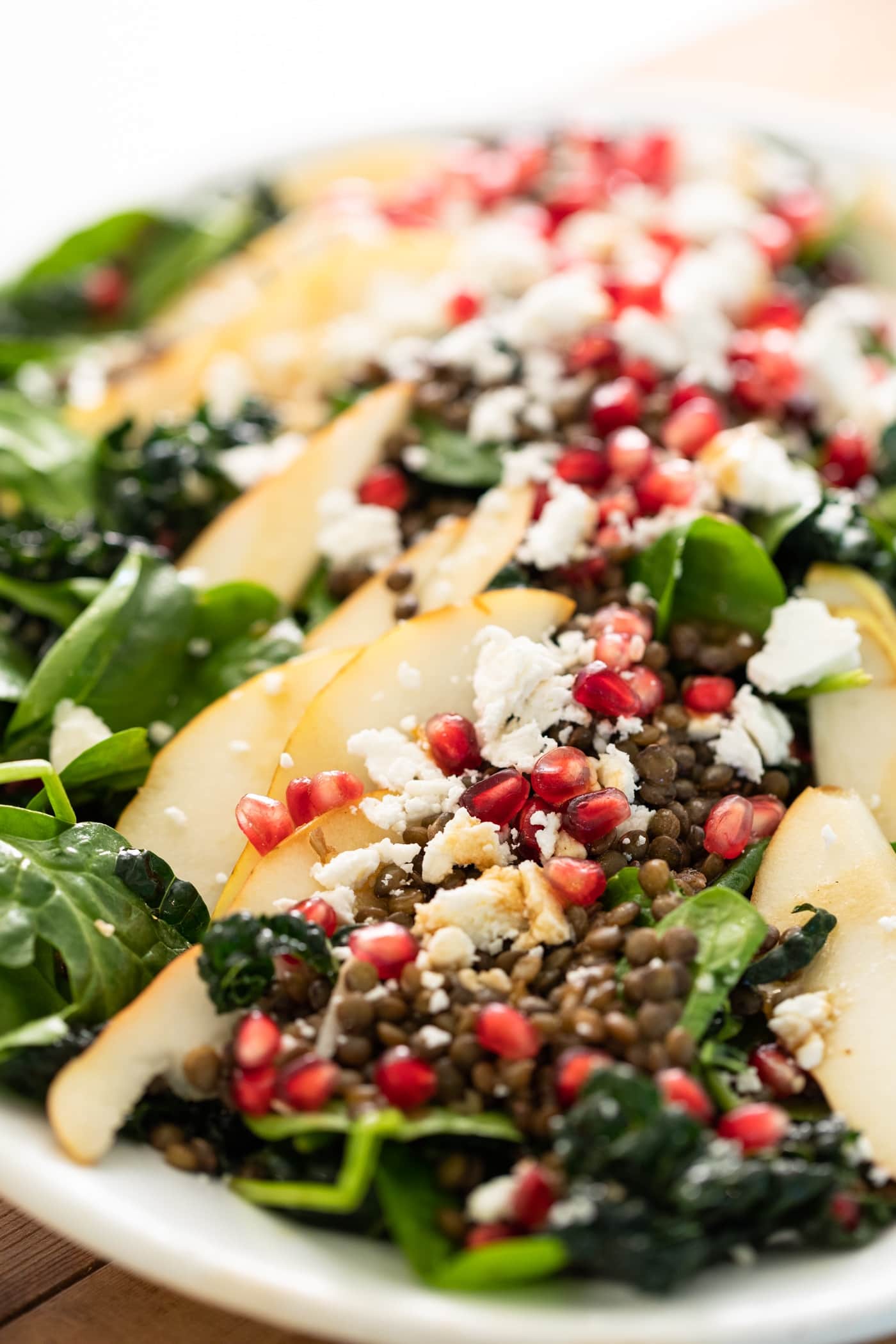 Close view of big slices of pear laying on top of green kale and spinach and topped with pomegranate seeds and white crumbles of feta cheese