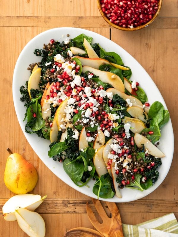 Top down view of large white platter filled with kale and spinach leaves and topped with pears, lentils, pomegranate arils, and crumbles of white feta cheese