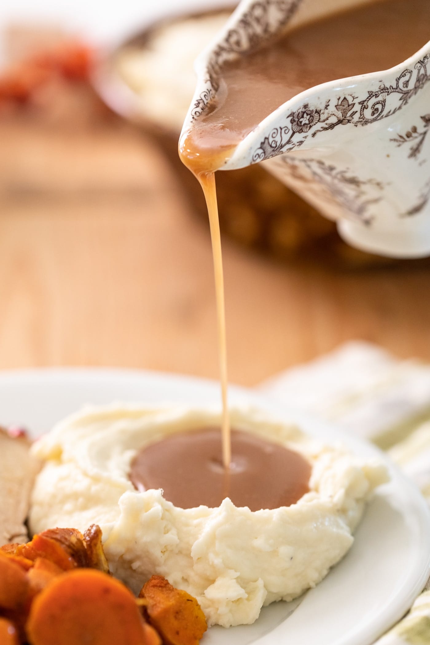 Stream of brown colored gravy being poured out of gravy boat onto a pile of white mashed potatoes with carrots and turkey on plate