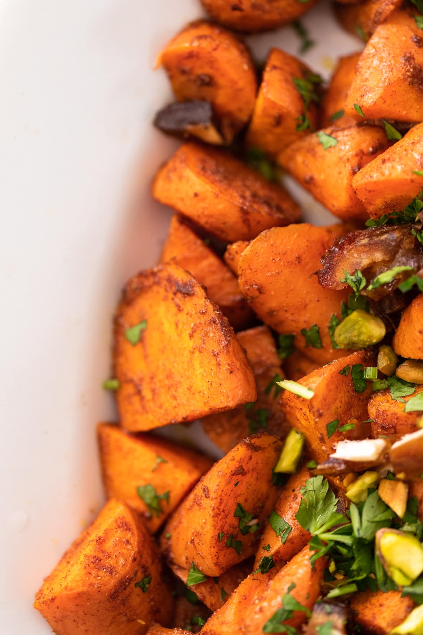 Orange carrots tossed in spice blend with brown flakes sitting in white bowl with bits of dates and carrots on top