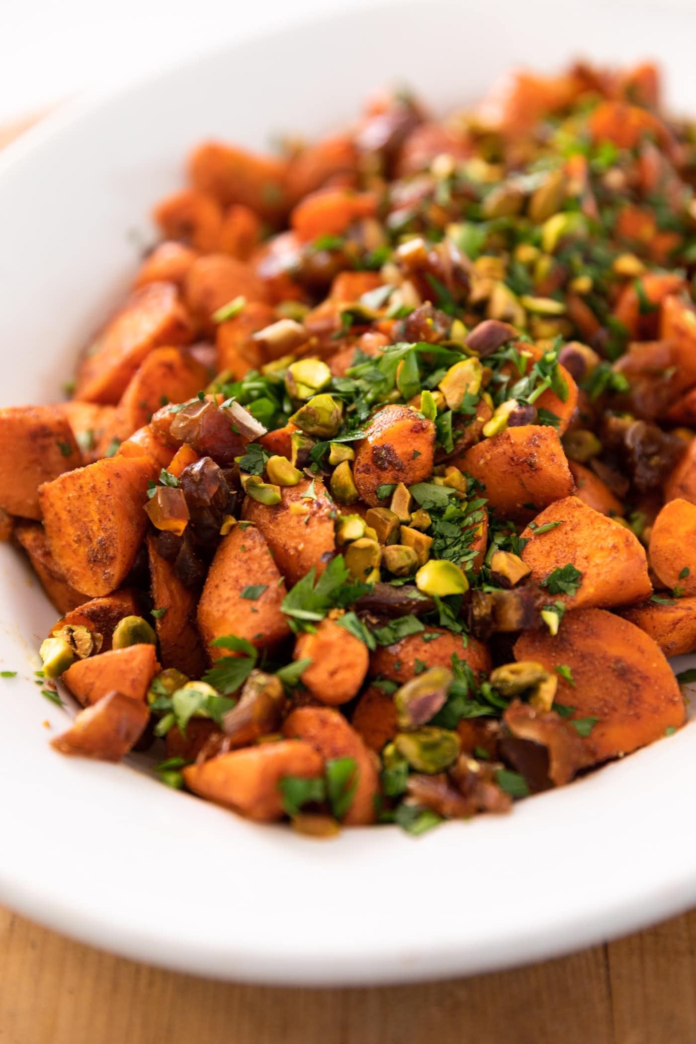 Roasted carrots piled into a white serving bowl topped with parsley, pistachios, and dates