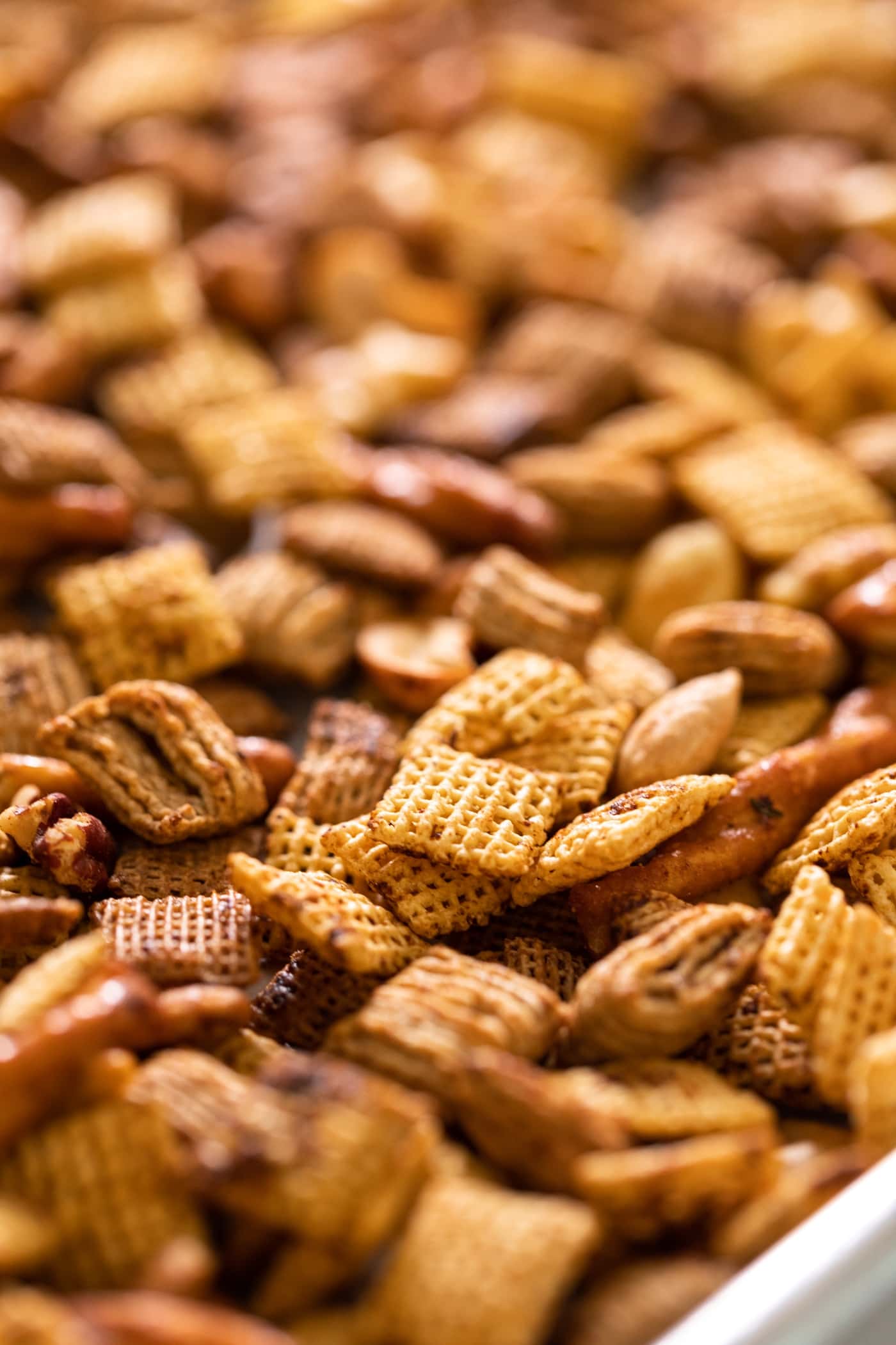 Close up view of piece of Corn Chex cereal sitting on top of other pieces of cereal and pretzels, all browned in the oven