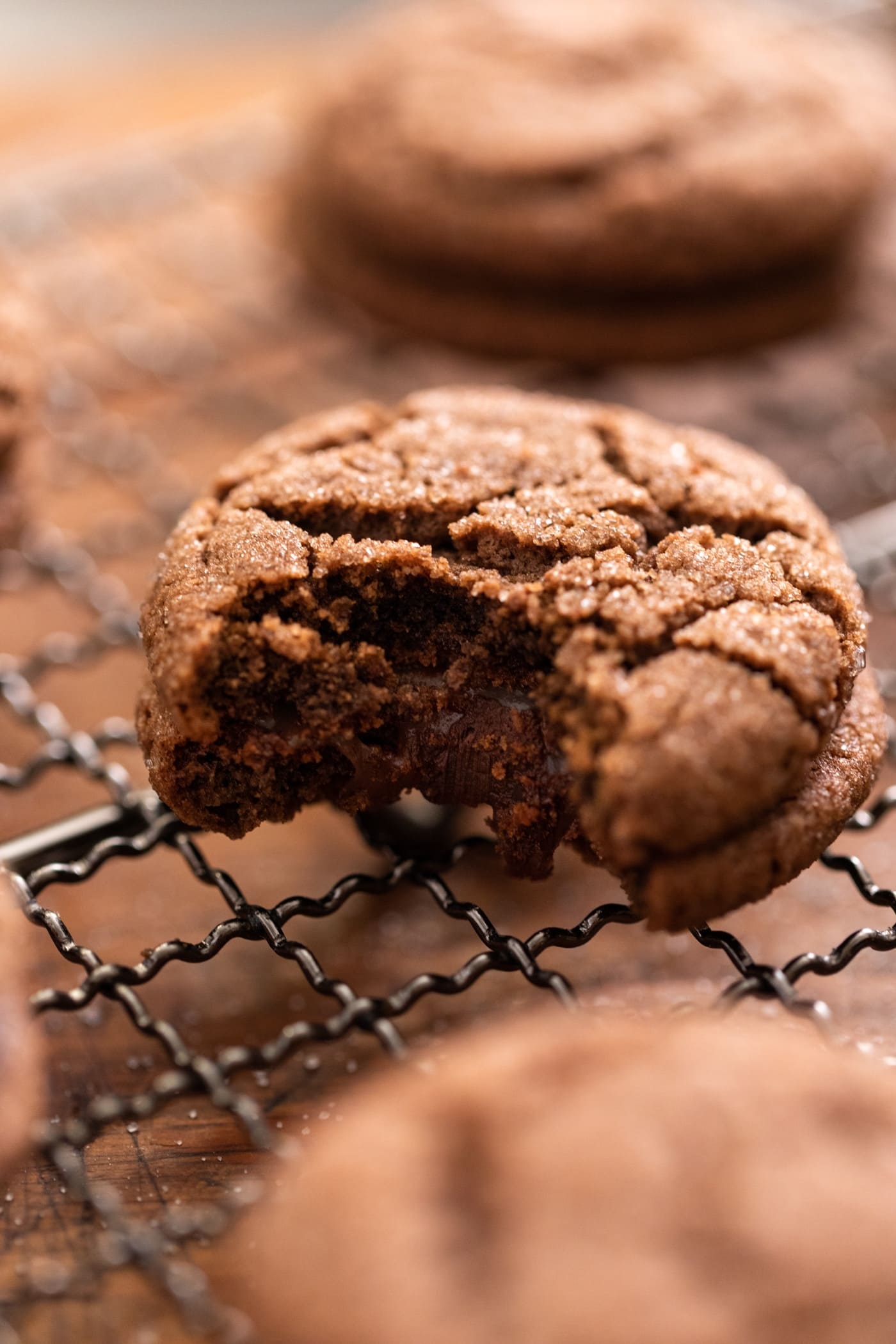 Chocolate cookie with bite taken out showing inside with gooey chocolate filling sitting on cooling rack with other chocolate cookies all around