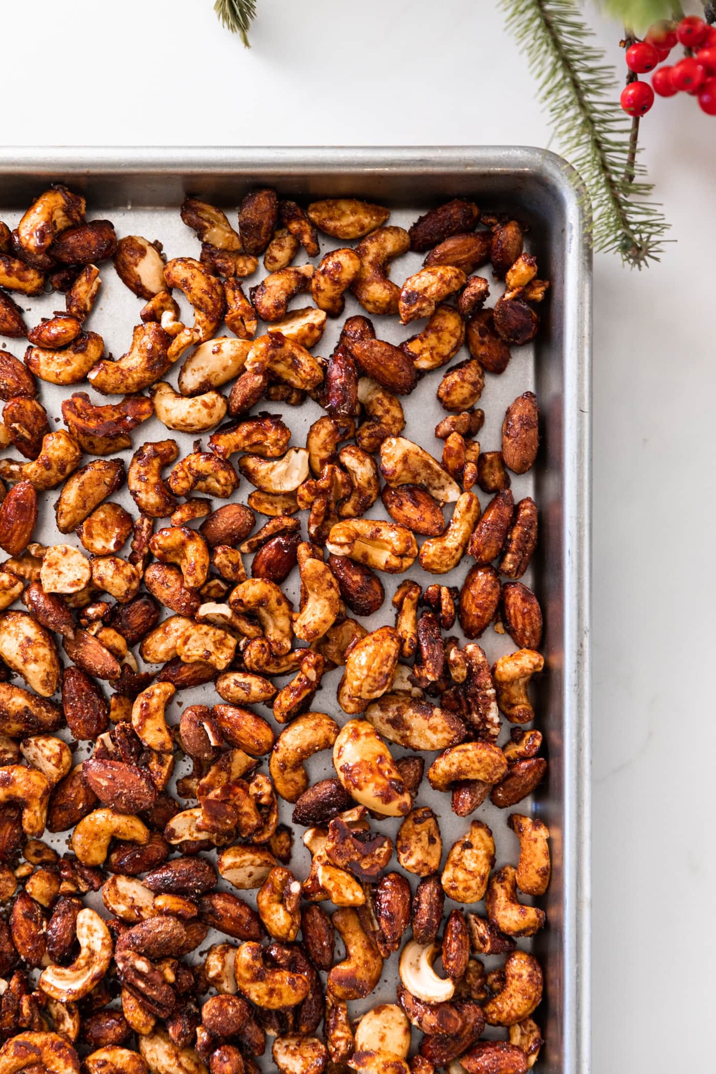 Top down view of cinnamon spiced nuts sitting on silver baking pan on white countertop with Christmas greenery and red berries to the side