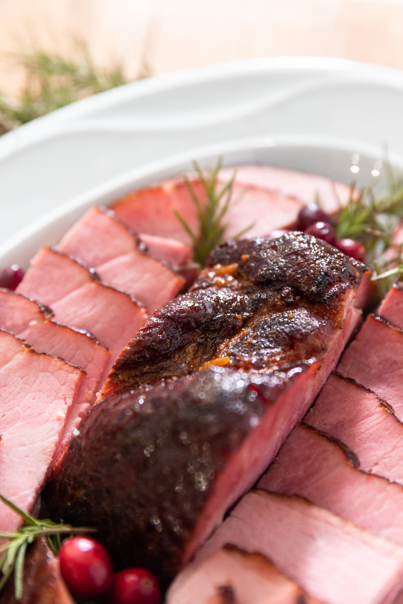 Piece of ham sitting on white platter surrounded by slices of ham along with sprigs of rosemary and red cranberries