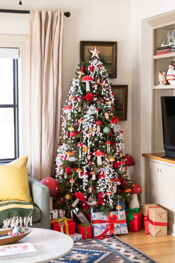 Large Christmas tree packed with red colored mushrooms and snow garland surrounded by presents on the floor and TV cabinet beside