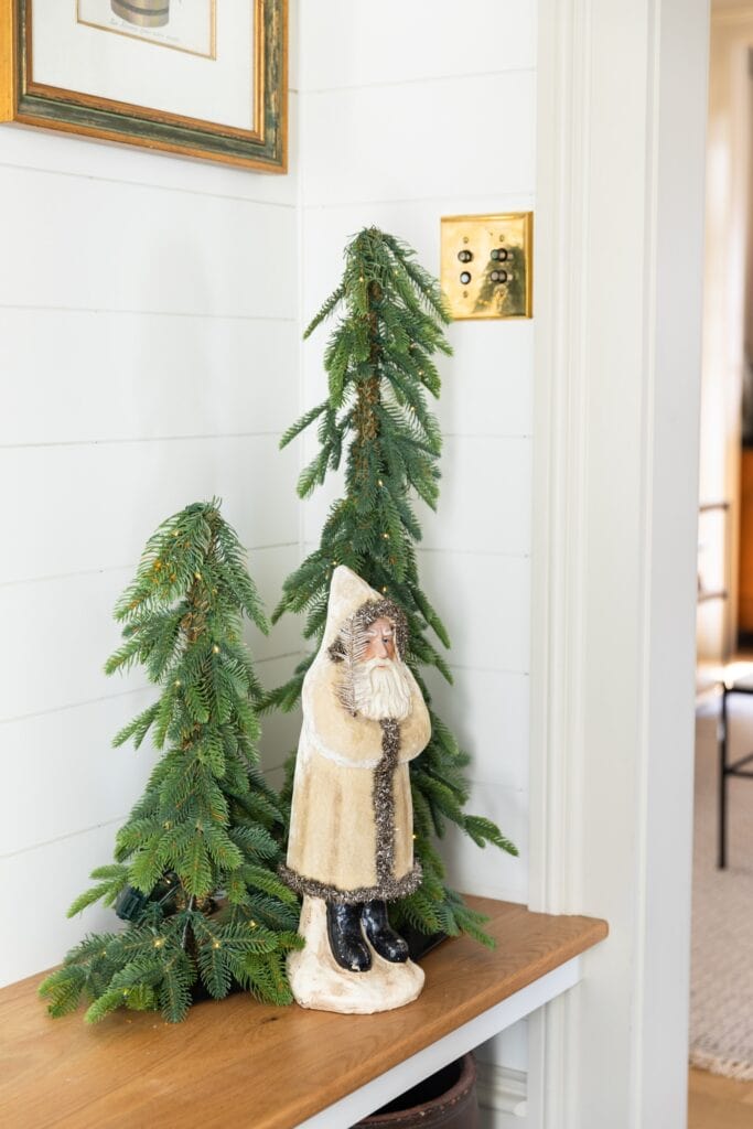 Small white coated toy Santa standing on wooden bench with two green trees with lights right behind