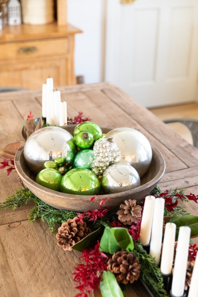 Wooden basket on wooden table filled with green and silver glass Christmas ornaments with greenery and candles all around
