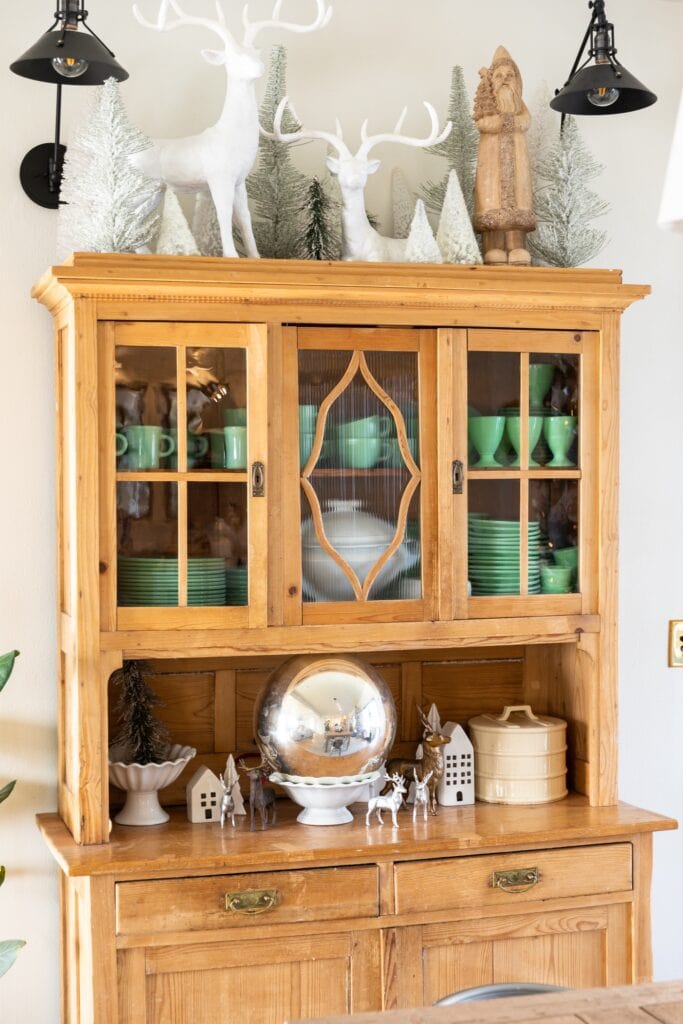 Light pine colored cabinet filled with green mugs and plates topped with white deer and Christmas trees
