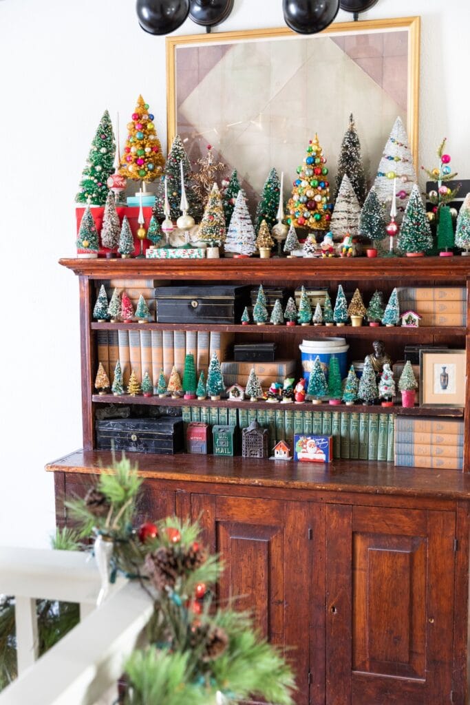 Dark wood cabinet piled with all sizes of Christmas bottle brush trees and small figures with a painting and spot lights above