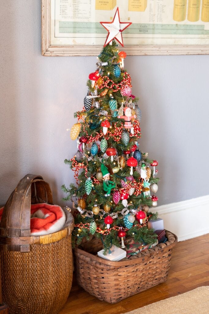 Half size Christmas tree covered with brightly colored vintage glass ornaments sitting in a basket with small vintage boxes piled all around the base