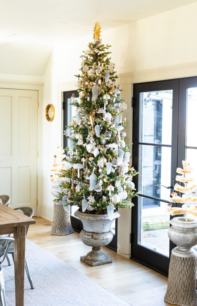 Large Christmas tree in concrete urn sitting in dining room covered in golds and silvers with black French doors behind leading onto patio