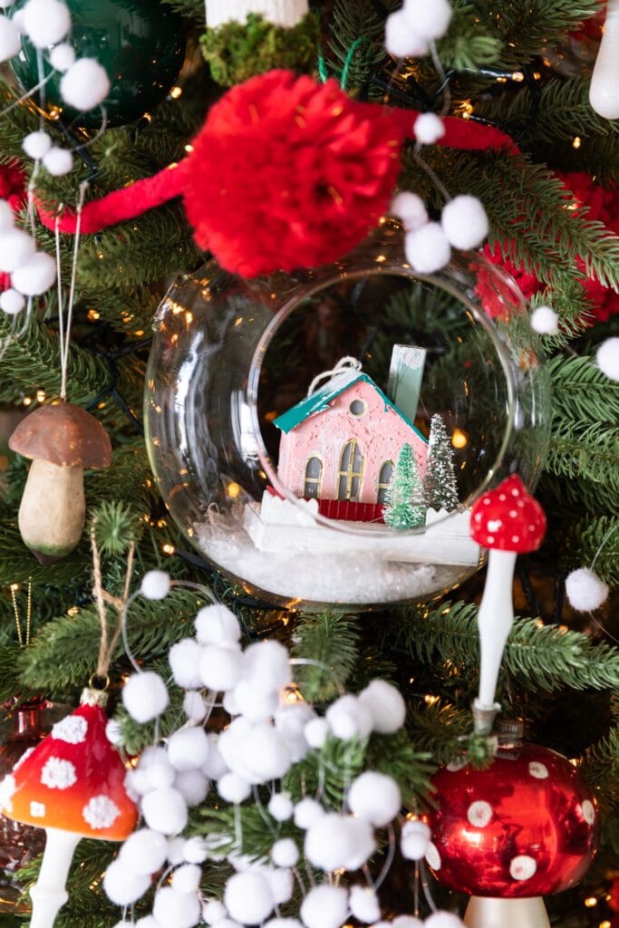 Close up view of glass dome with small pink paper house inside surrounded by mushroom ornaments on a Christmas tree