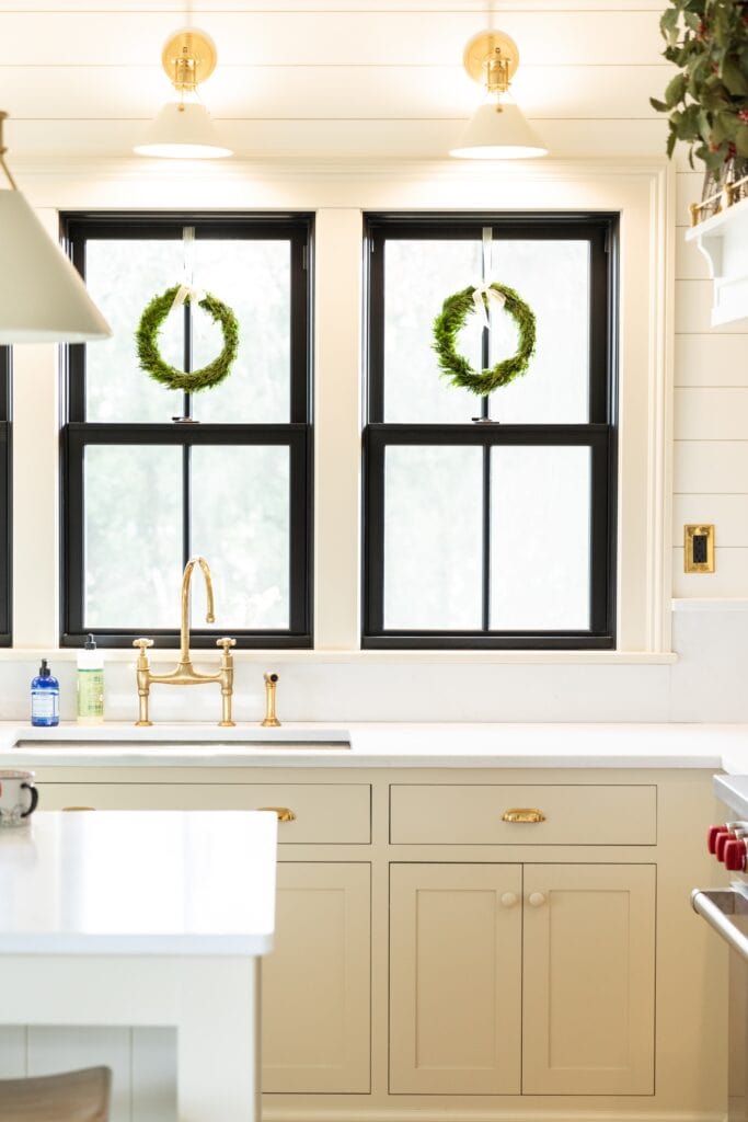 Two windows with green wreaths hanging from tops in white kitchen with white countertops and off-white cabinetry