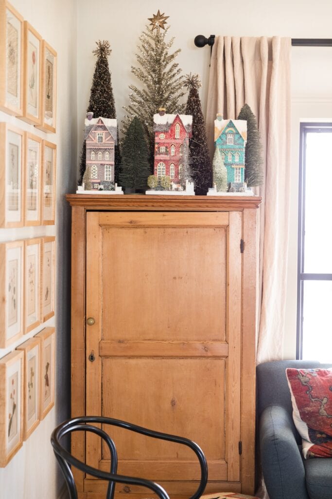 Pine chest topped with tall Christmas houses along with trees behind with couch to the side and frames on the wall on the other