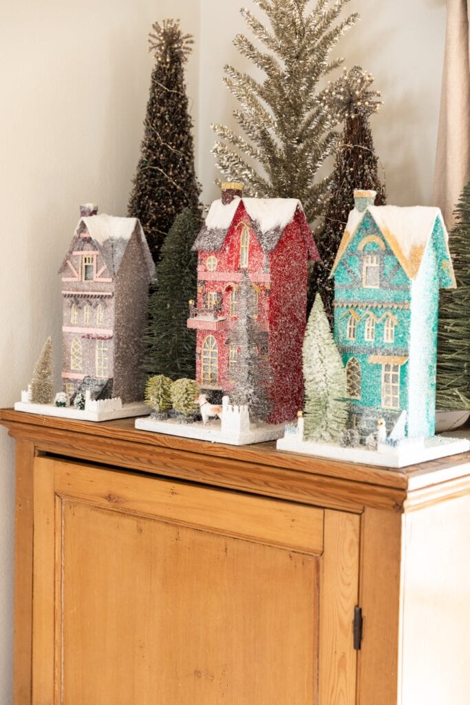 Three tall display Christmas houses with yards covered in snow sitting on top of pine dresser with trees in background