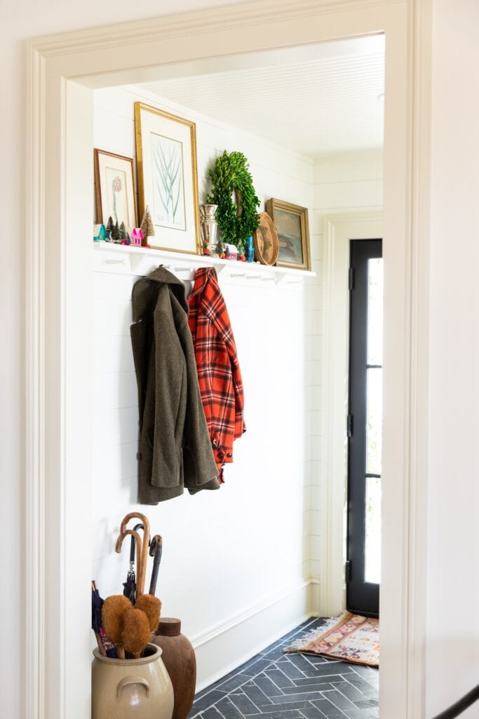 Looking into an entryway with black tile floor with pegs on the wall and coats hanging up with shelves adorned with Christmas decor