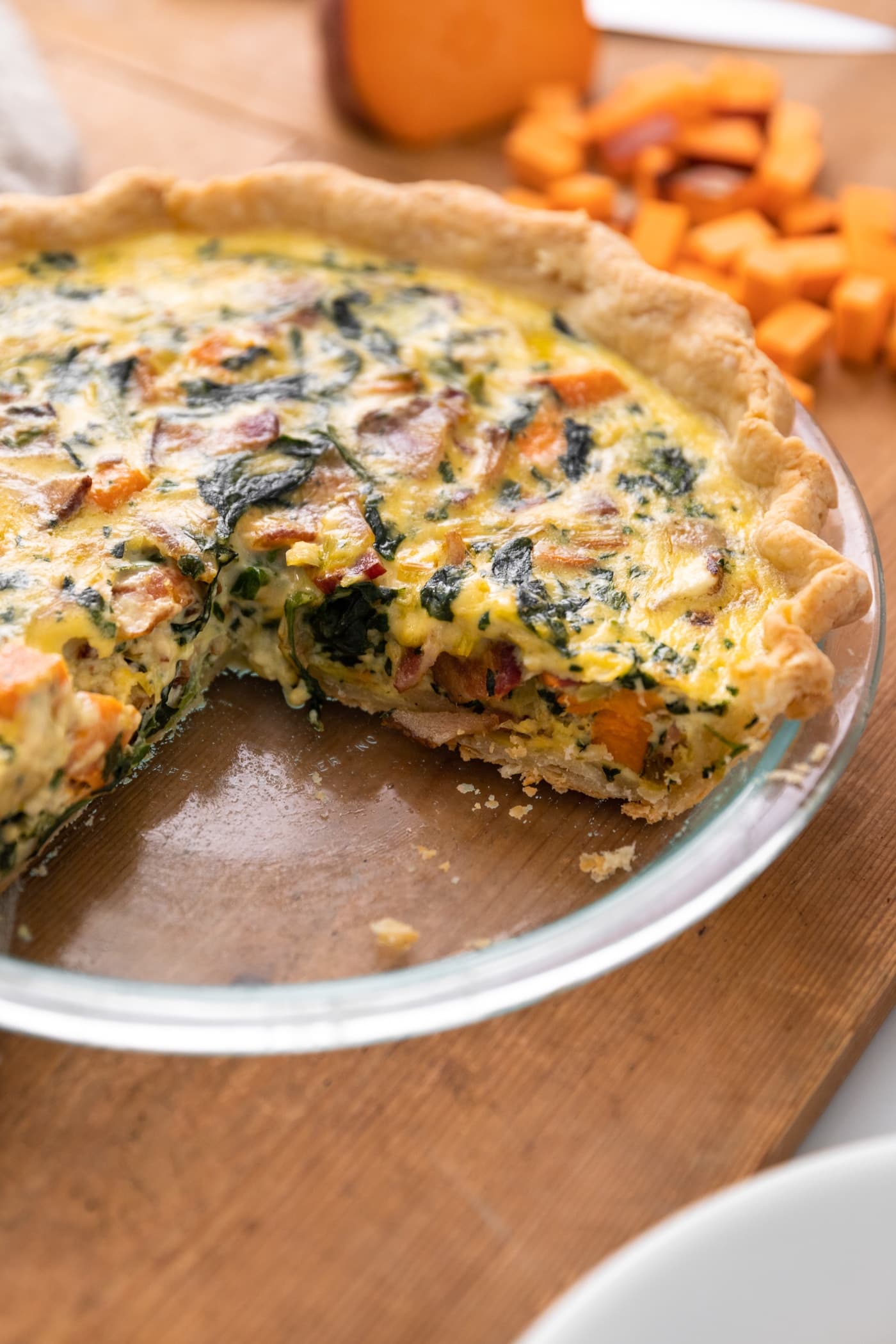 Glass pie plate containing yellow quiche with several pieces cut out with extra chunks of sweet potato in the background all on wooden surface