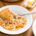 White plate filled with white beans, ham, and carrots with piece of cornbread in amongst soup all on wood board