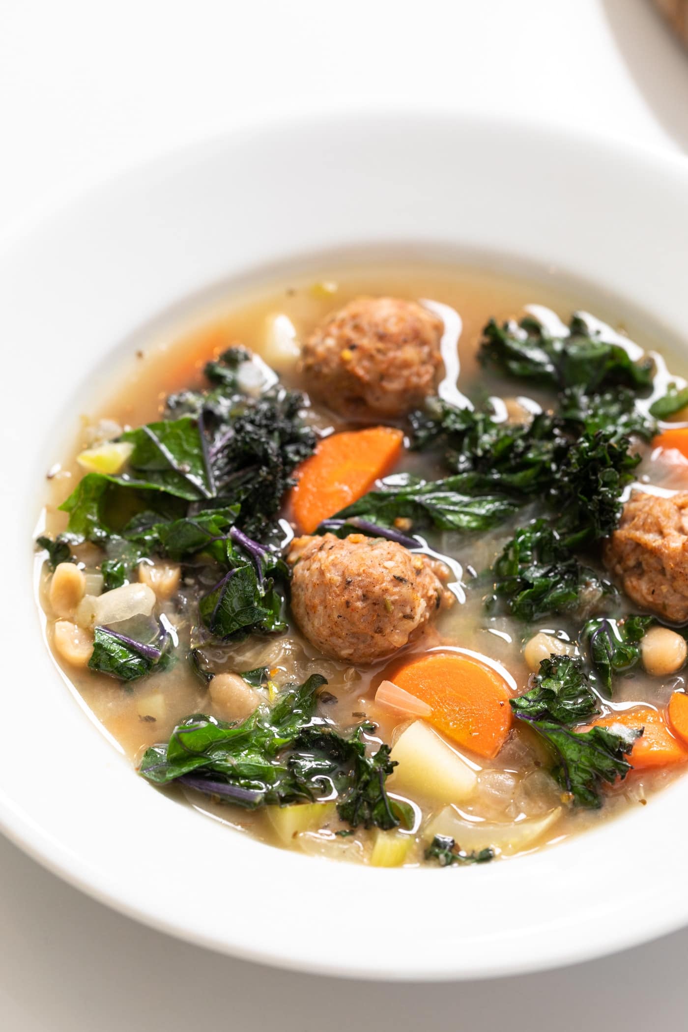 White bowl sitting on marble countertop filled with broth soup filled with meatballs and carrots and pieces of kale