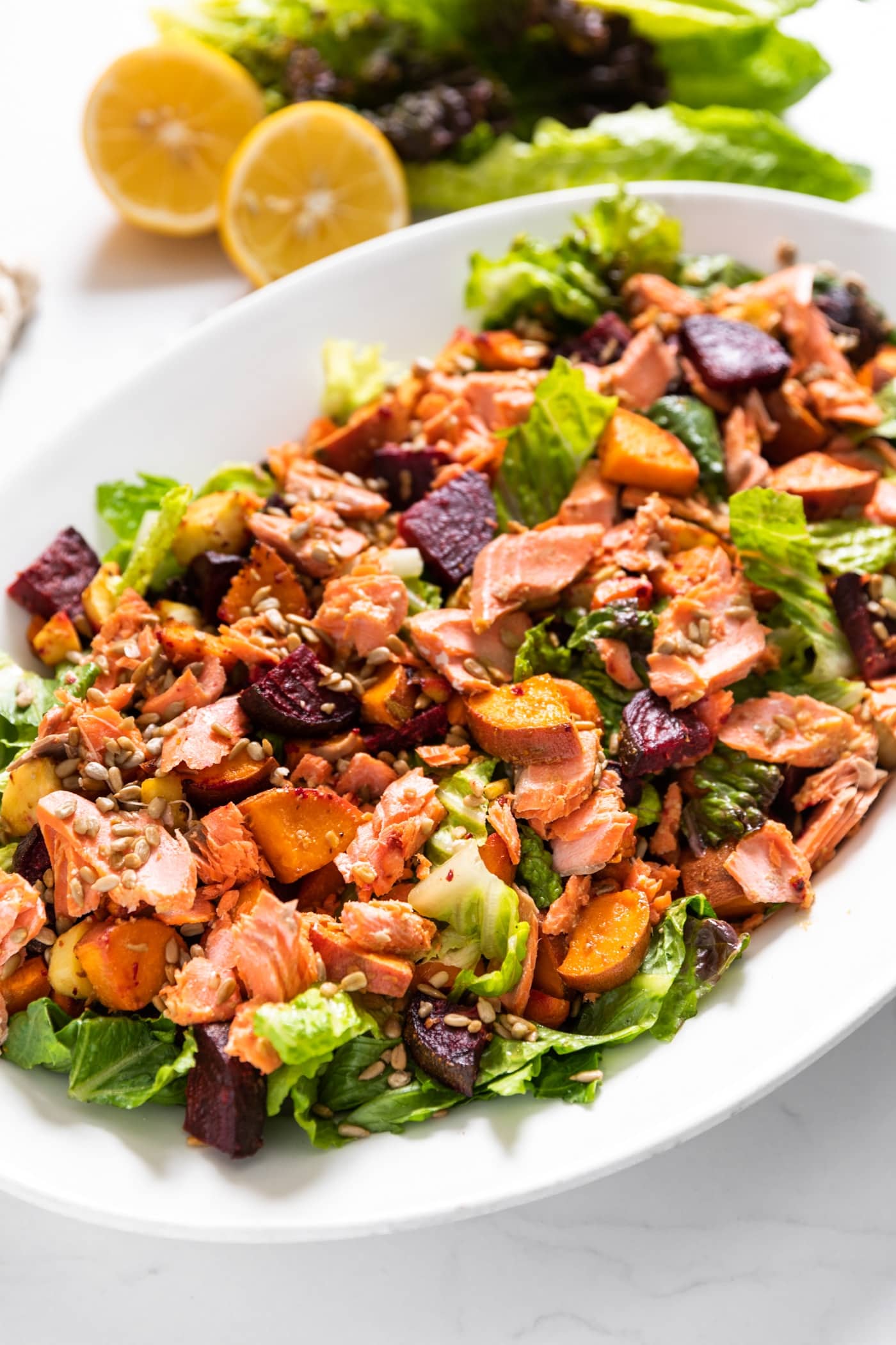 Large white platter sitting on white countertop filled with salad greens topped with beets and carrots and sweet potatoes and salmon with cut lemons in background
