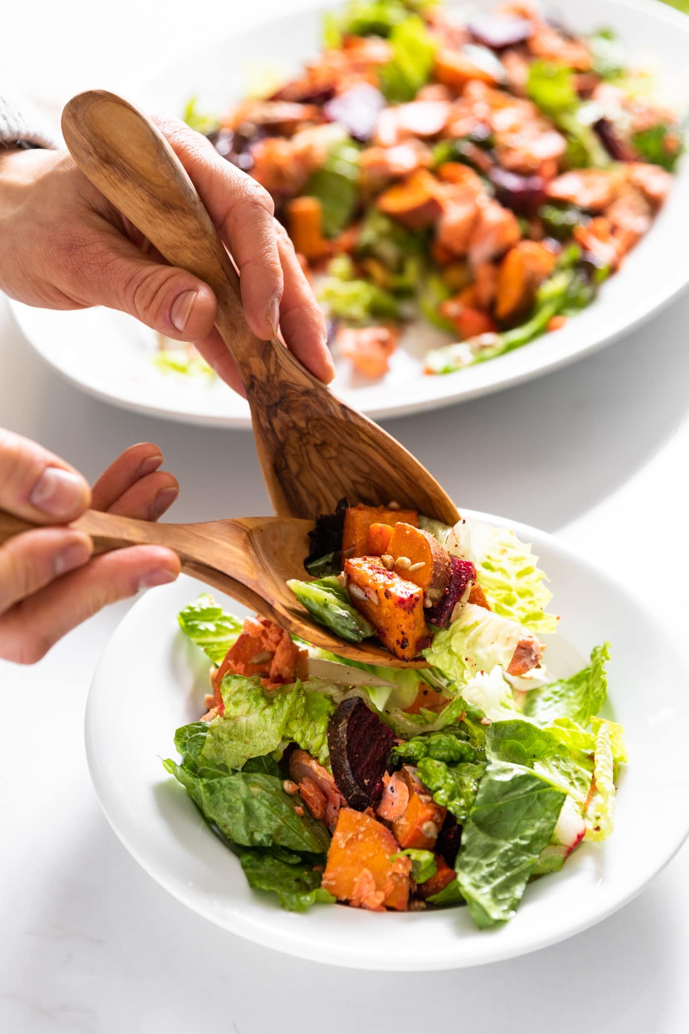 Two hands holding tongs spoon salad onto white plate from salad sitting in white plate behind