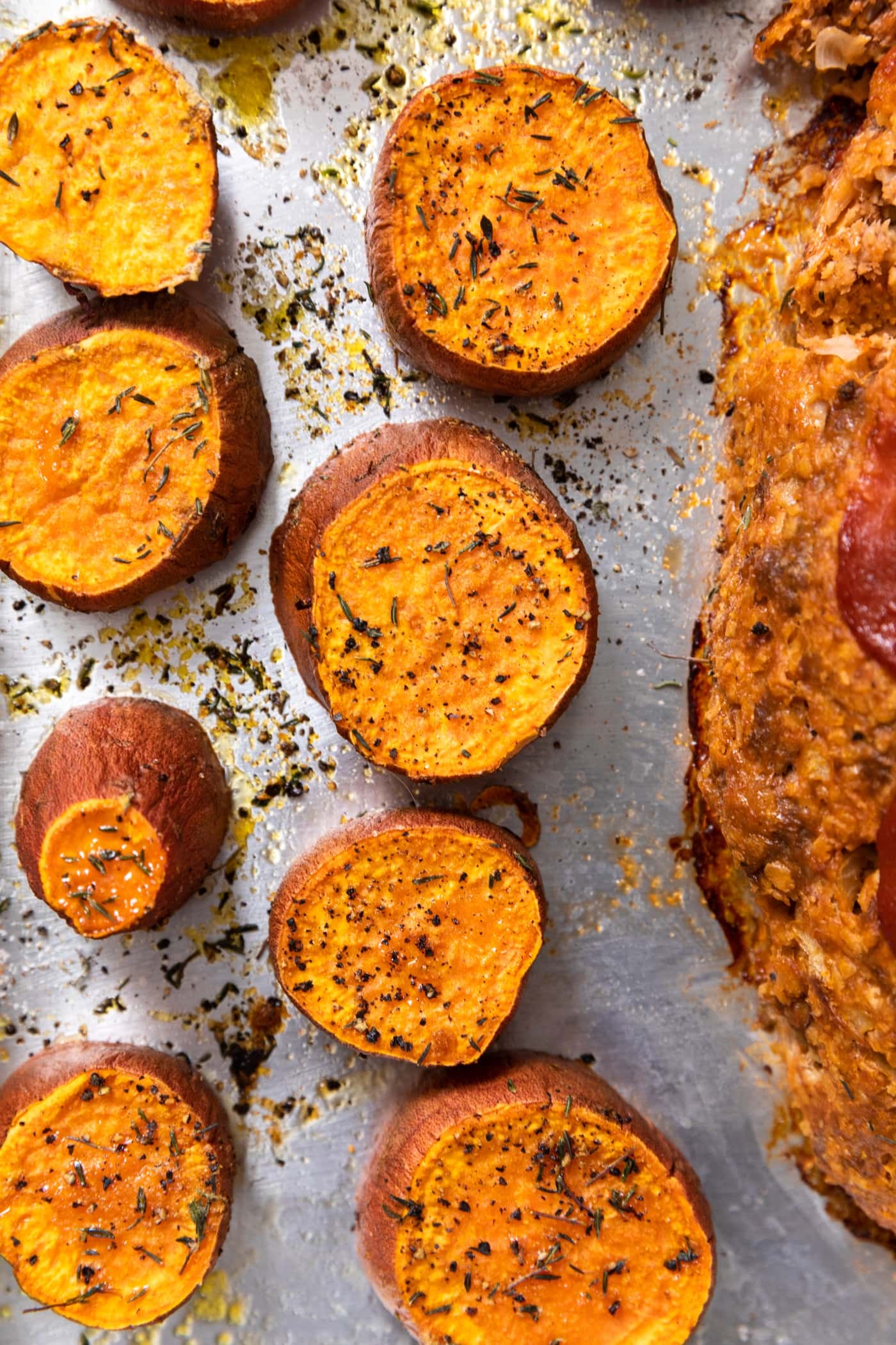 Slices of orange colored sweet potato sitting on stainless steel sheet pan sprinkled with bits of herbs