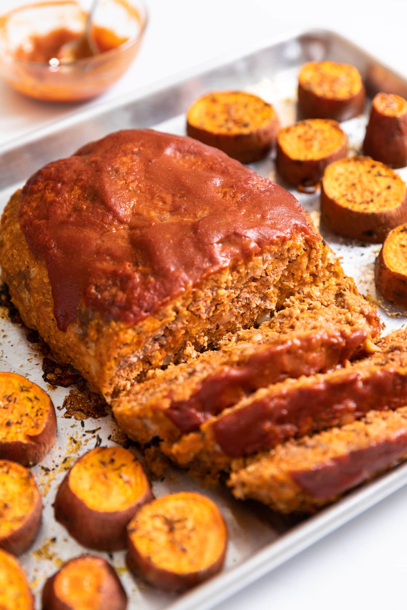 Meatloaf covered with red ketchup on top with slices laying on sheet pan with orange colored sweet potatoes all around