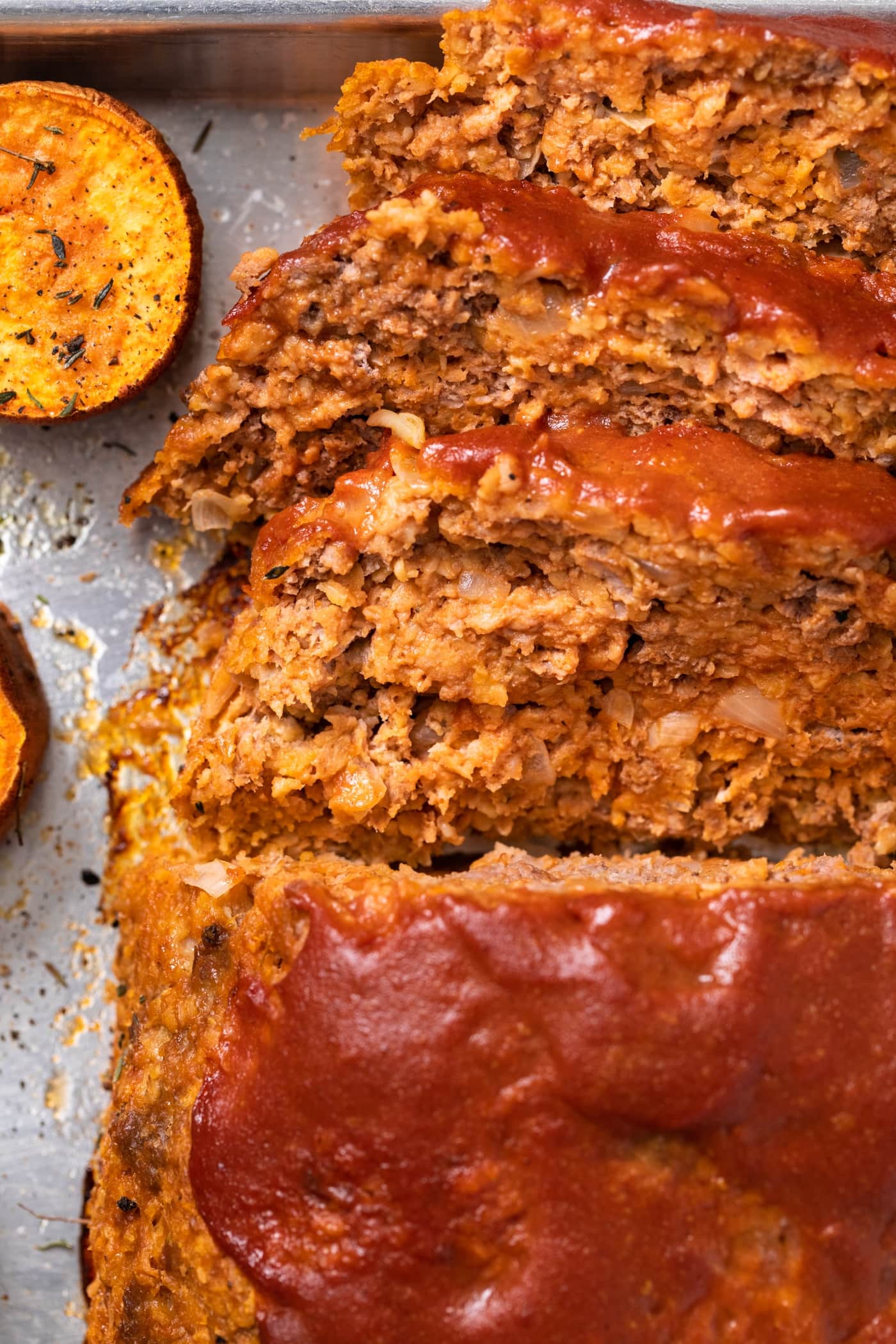 Close up top down view of slices of meatloaf laying on sides of sheet pan with one slice or orange colored sweet potato
