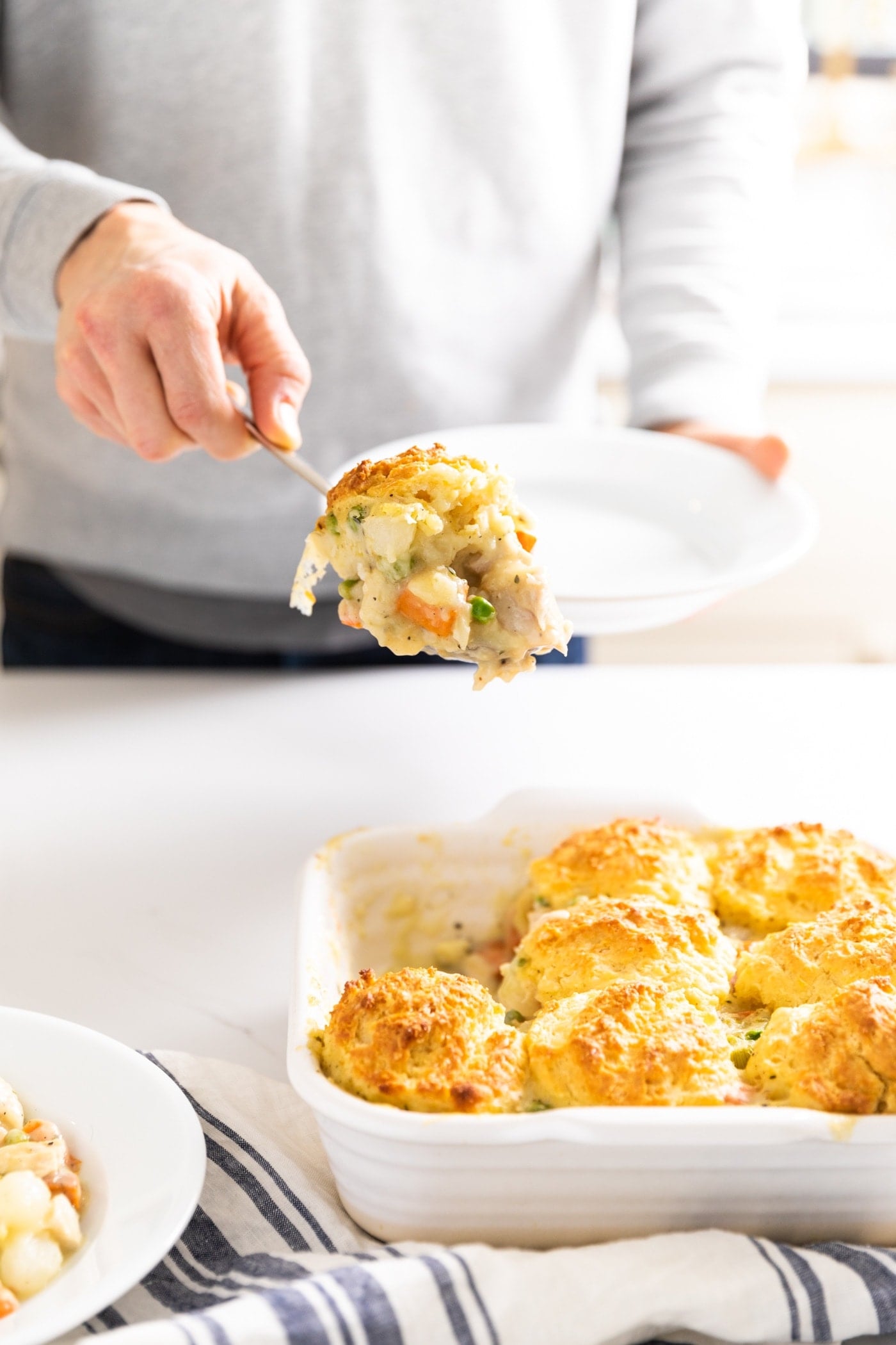 Hand holding scoop of chicken pot pie mixture with biscuit on top with container of biscuits underneath