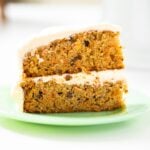 Slice of layered carrot cake sitting on green plate on a white countertop with a white cake plate in background