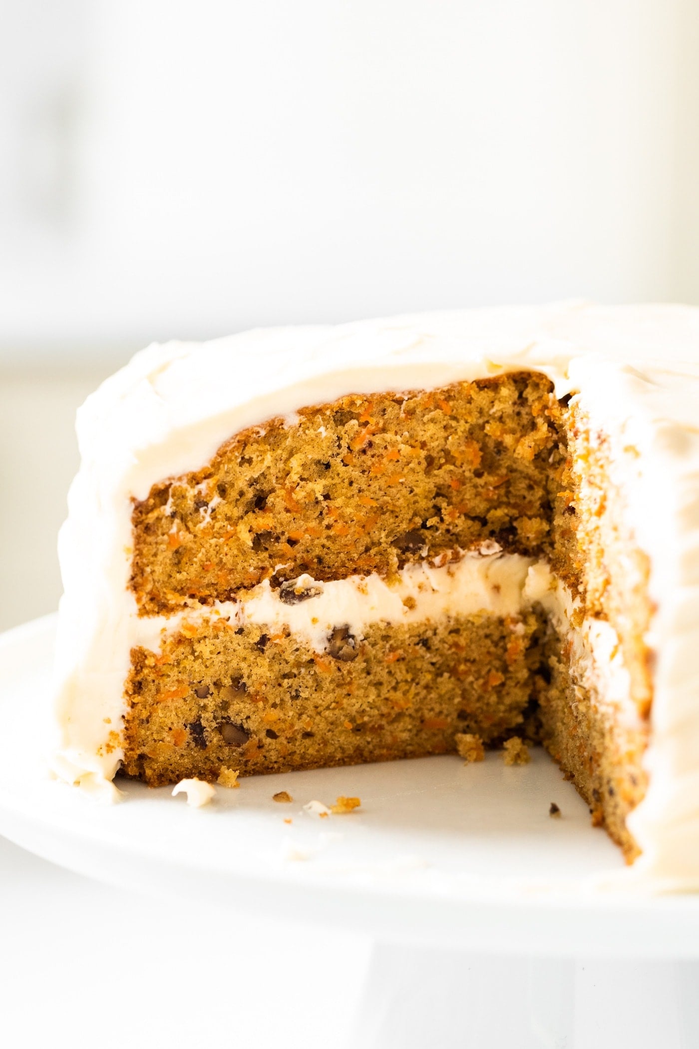 Close up view of inside of orange colored carrot cake topped with cream cheese frosting sitting on white cake plate with white background