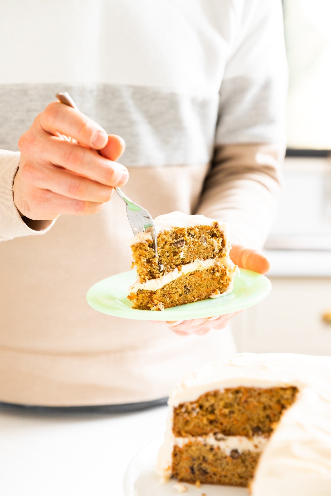 Hand holding green colored plate with slice of carrot cake with fork slicing into cake with rest of cake in front