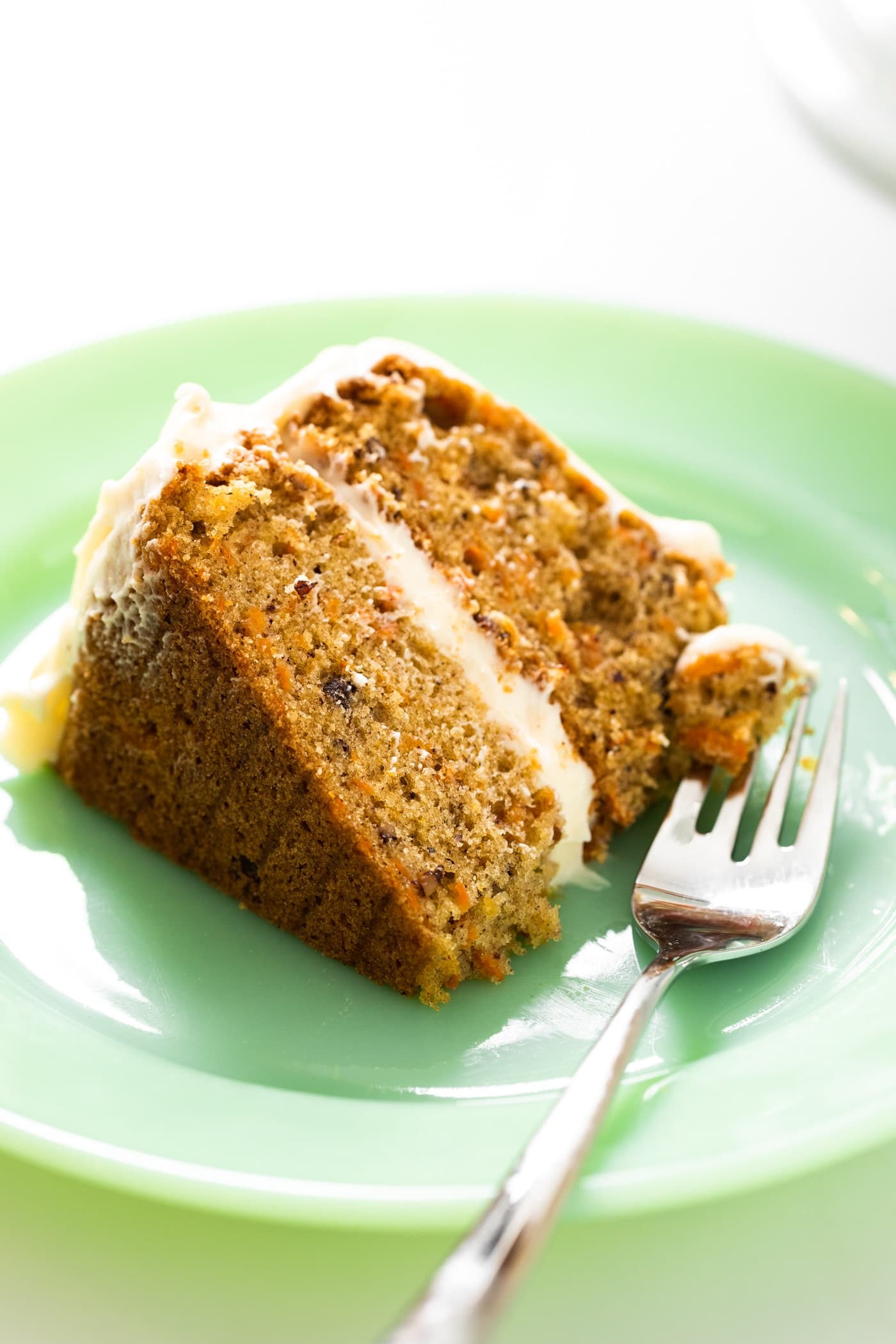 Big slice of carrot cake with two layers and white cream cheese frosting in between layers sitting on green colored place with fork