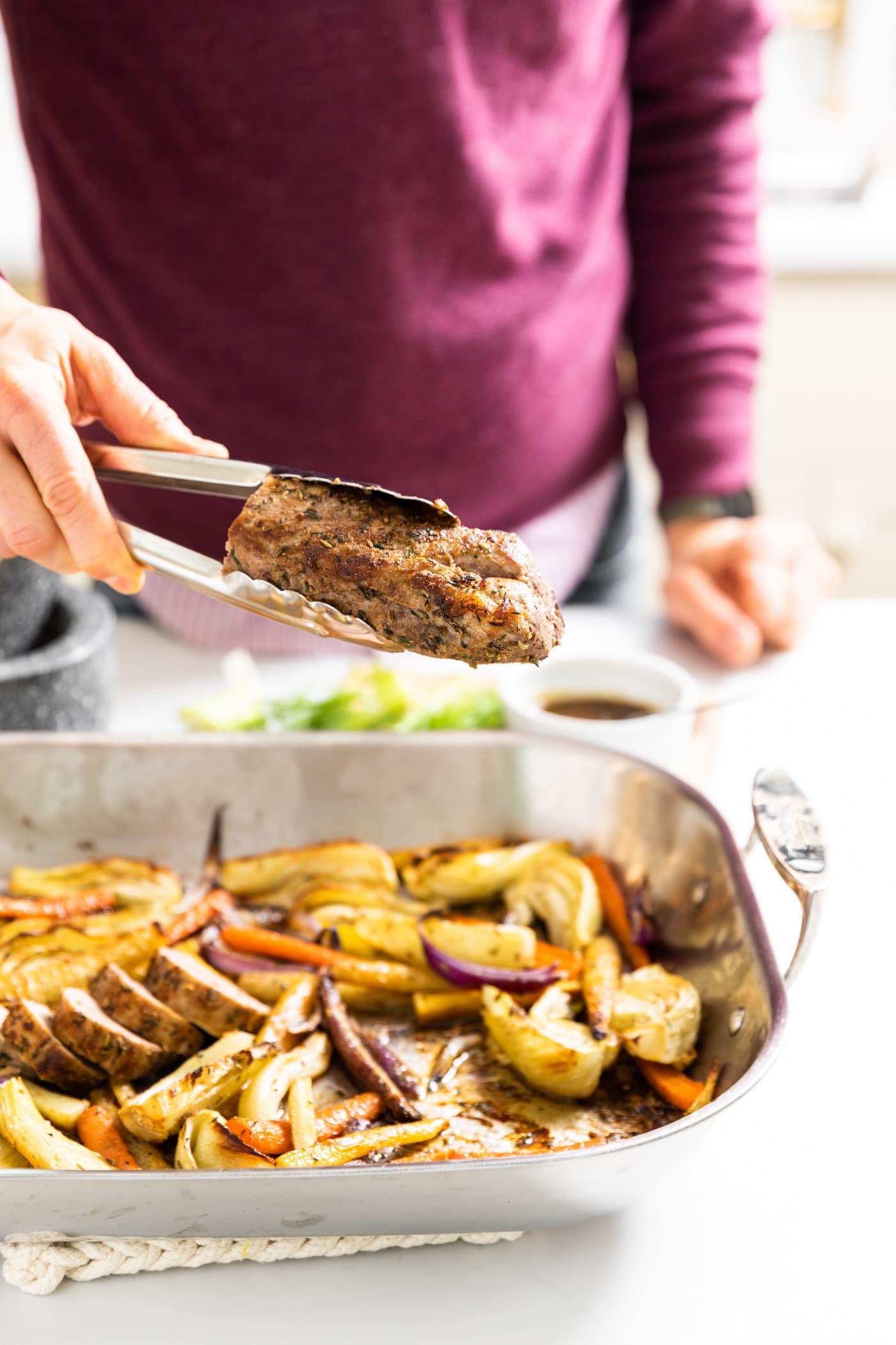 Hand holding tongs clutching a pork tenderloin roast with pan below it filled with slices of roasted vegetables all on white countertop